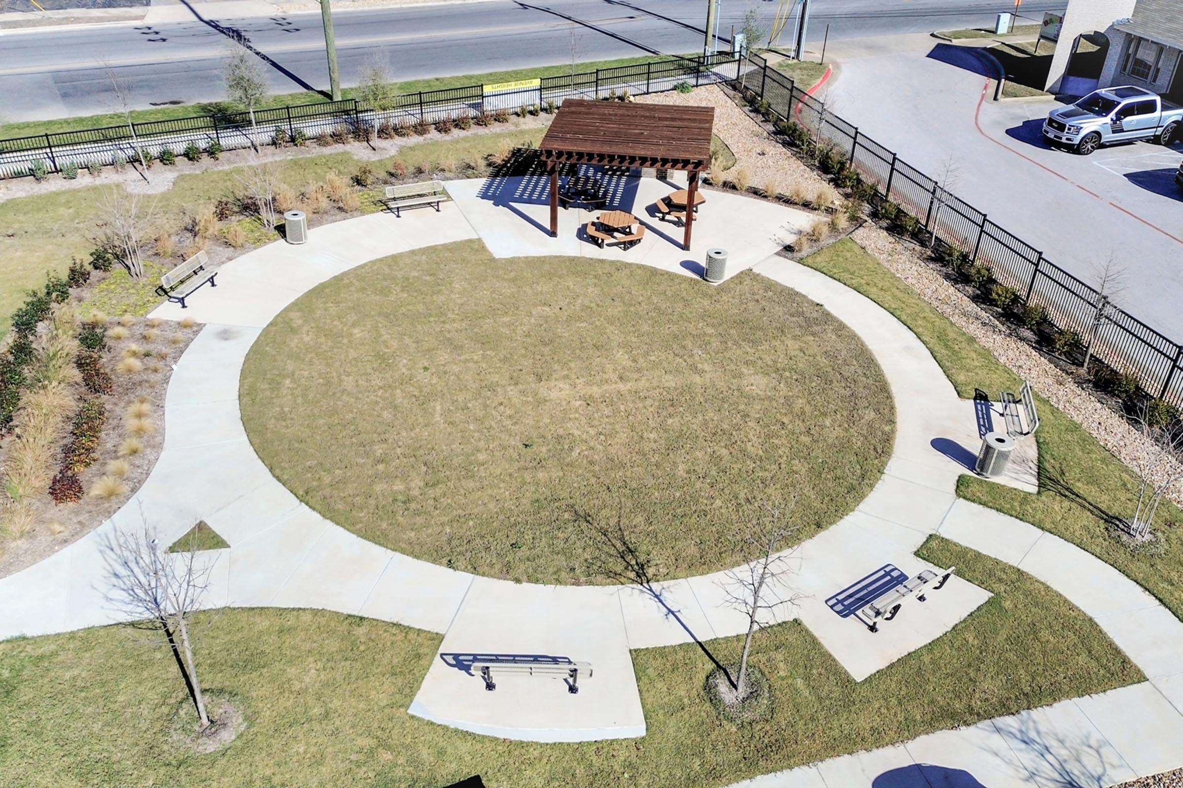 Aerial view of a circular park area featuring a gazebo with tables, surrounded by green grass and pathways. Benches are placed along the perimeter, with a fence and road visible in the background. The park appears well-maintained, suitable for leisure activities and gatherings.