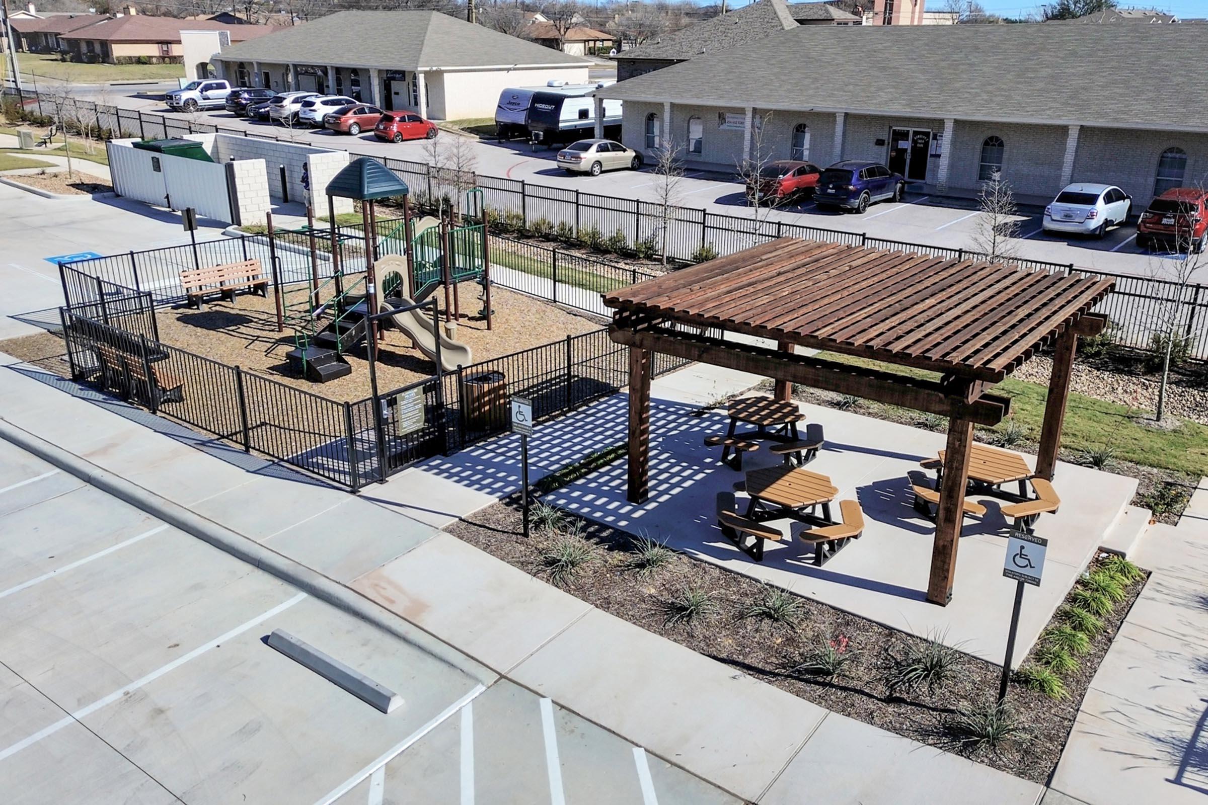 Aerial view of a playground area with a wooden structure and picnic tables. The playground features a climbing structure and swings, surrounded by a safety fence. Nearby, there is a parking lot with parked vehicles and landscaped greenery. The scene is bright and well-maintained, suitable for families.