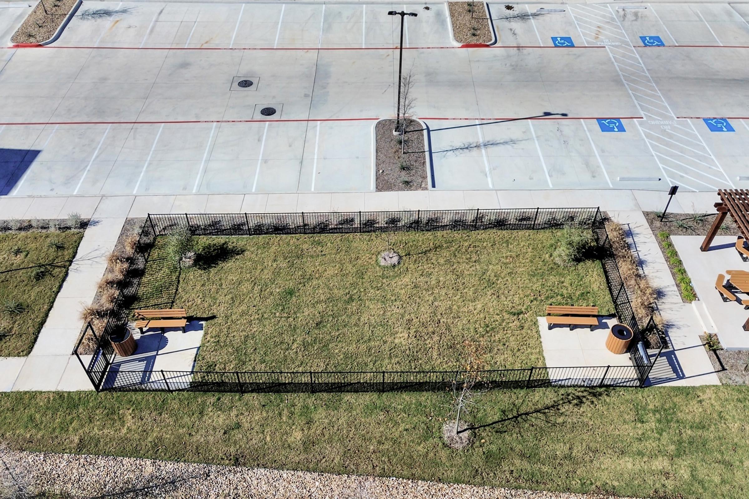 Aerial view of a fenced green space with two benches and decorative pots, surrounded by sidewalks. Nearby, a large empty parking lot with designated spaces. The area features manicured grass and sparse landscaping elements.