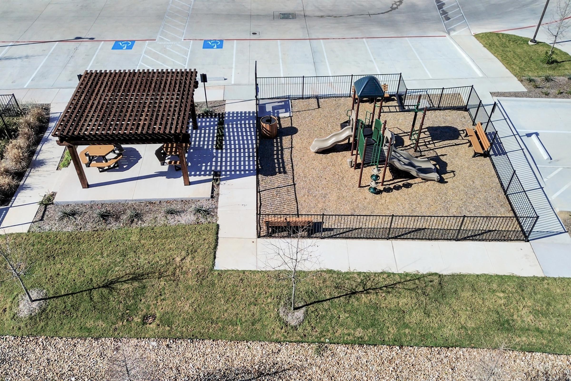 Aerial view of a playground area featuring two green slides, climbing structures, and benches under a wooden gazebo. The space is enclosed by a black fence, with a grassy area and a gravel surface surrounding the play equipment. Nearby, accessible parking spaces are visible.