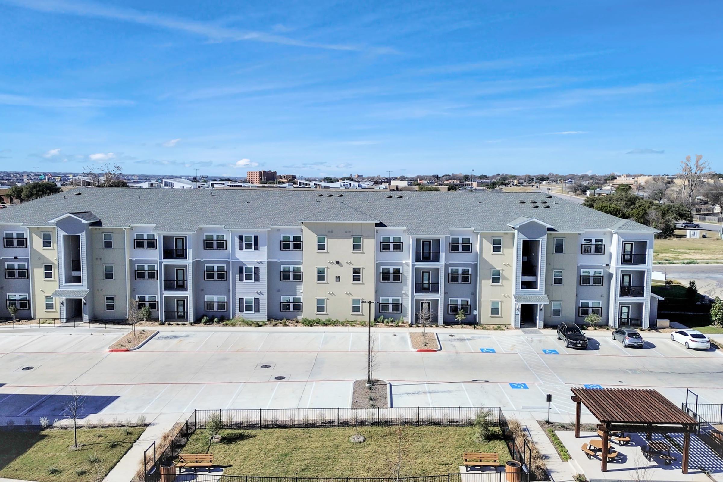 A view of a multi-story residential building with a mix of light blue and beige siding. The building has several balconies and windows. In front, there is a paved parking lot with parked cars and a grassy area with benches nearby. The sky is clear with a few clouds, and there are distant buildings on the horizon.