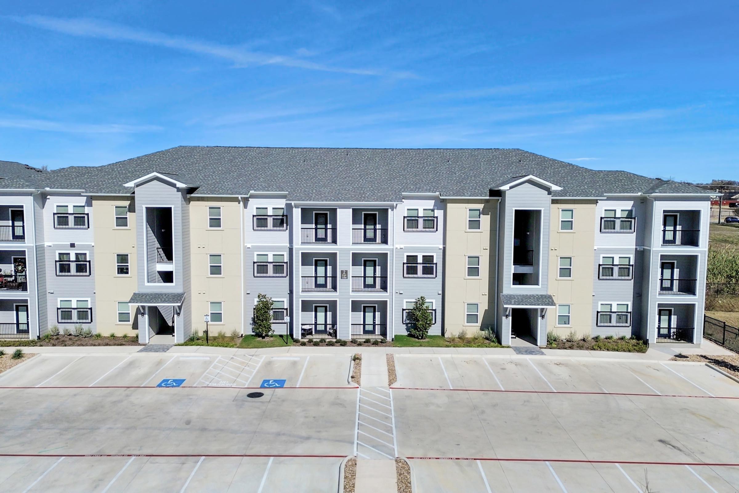 A modern multi-story apartment building with a light-colored exterior and dark accents. The building features multiple balconies and large windows. In the foreground, there is a paved parking lot with designated handicapped spaces. Clear blue sky provides a bright backdrop.