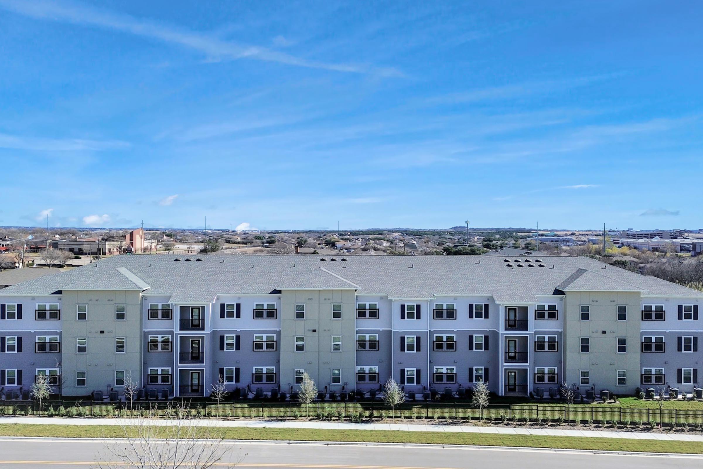 A row of modern multi-story apartment buildings with light-colored exteriors, surrounded by a landscaped area. The sky is bright blue with a few clouds, and there are mountains or hills in the background, indicating a suburban setting.