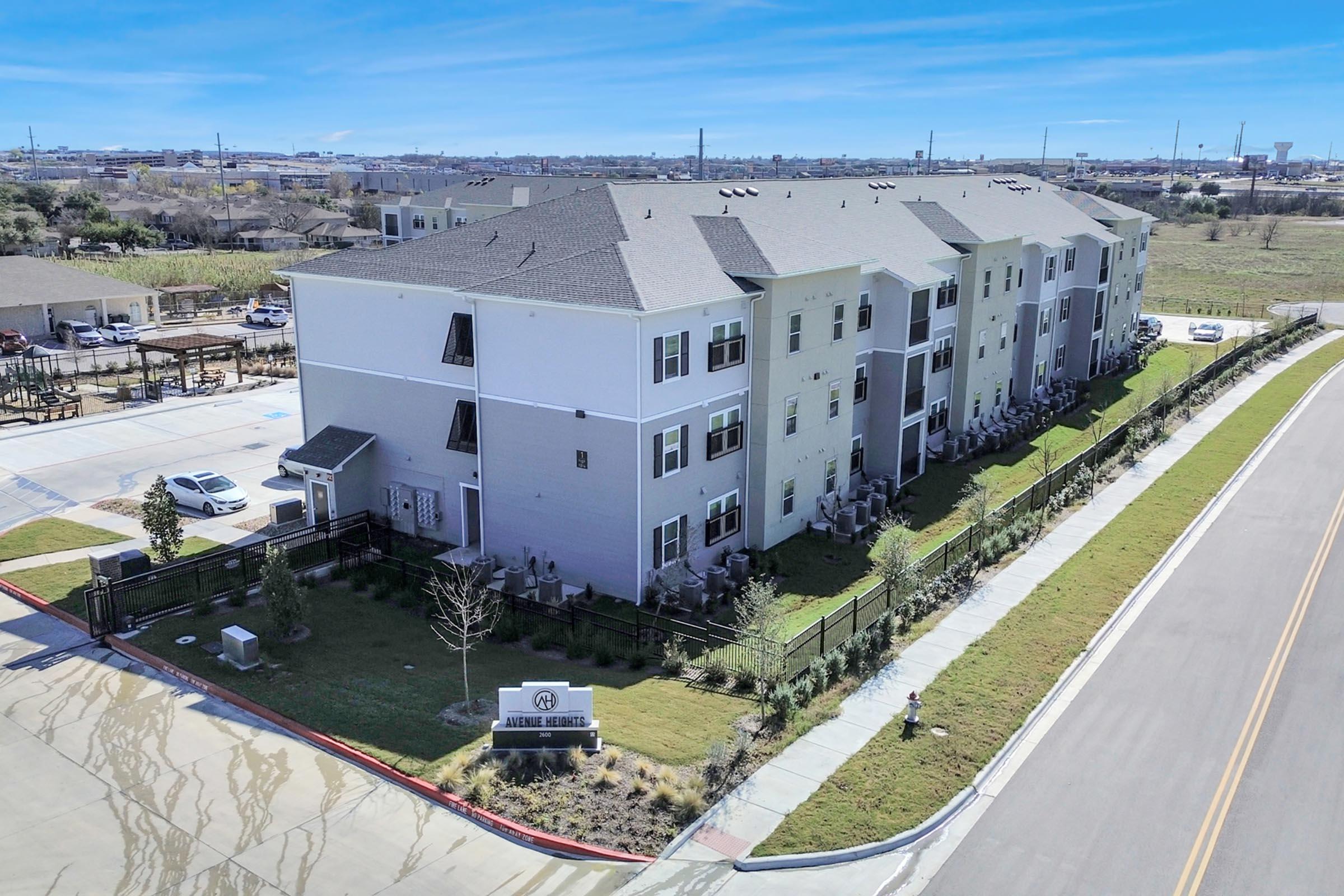 Aerial view of a modern residential building with multiple units. The building features a mix of gray and white exterior colors, surrounded by landscaped greenery and a sidewalk. In the foreground, there is a sign indicating the property name. The area appears well-maintained and includes a parking lot in the distance.