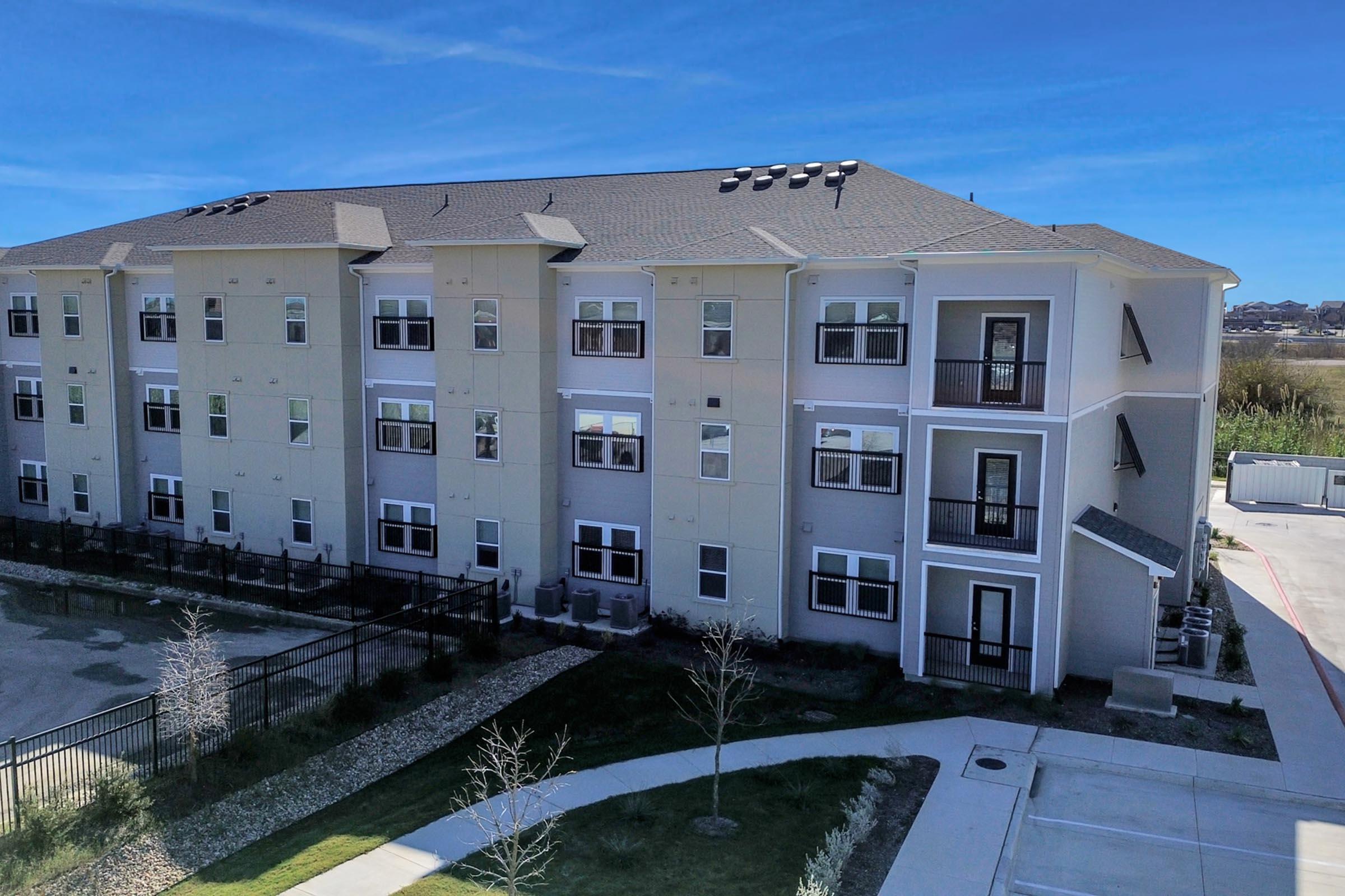 A modern multi-story apartment building with a light gray façade, featuring multiple balconies and large windows. The surrounding area includes landscaped greenery and a paved walkway, under a clear blue sky.