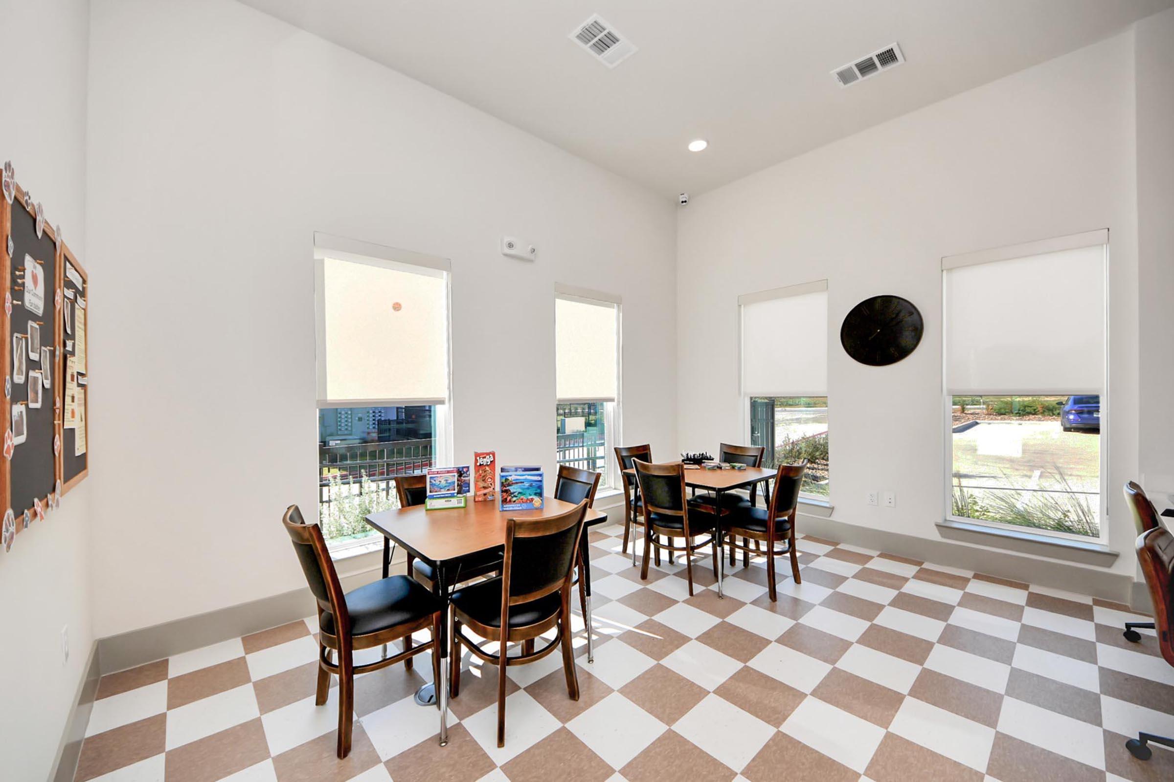 A bright and spacious room with a checkered floor, featuring two tables with chairs, large windows with shades, and a wall clock. There are some magazines or brochures on the tables, and a bulletin board on one wall, creating an inviting space for casual meetings or gatherings.