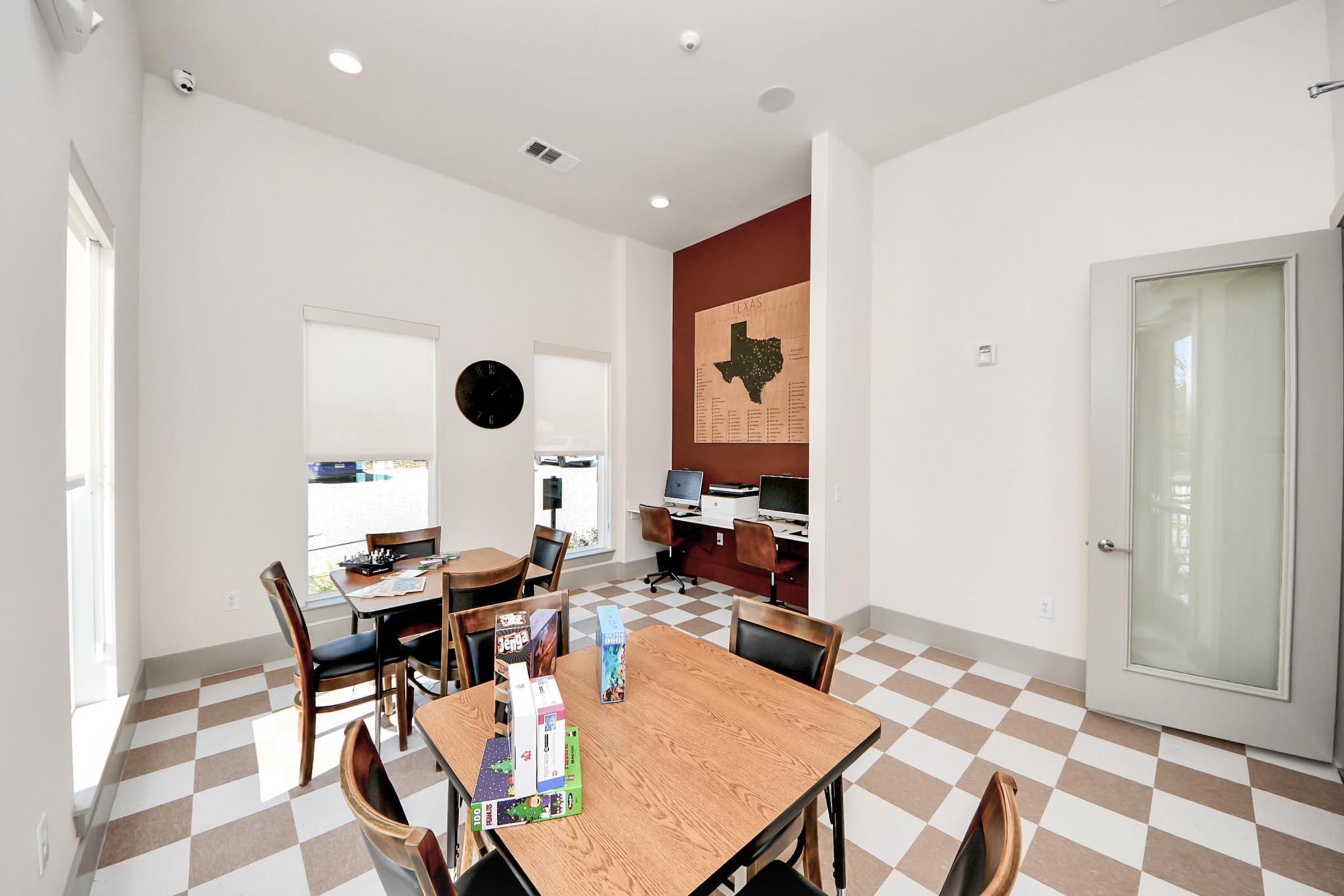 A bright and spacious room with a large wooden table surrounded by chairs. The walls are painted in light colors, with one wall featuring a Texas map art installation. There are two computers on a small desk and large windows letting in natural light. The floor has a checkered pattern of light and dark tiles.