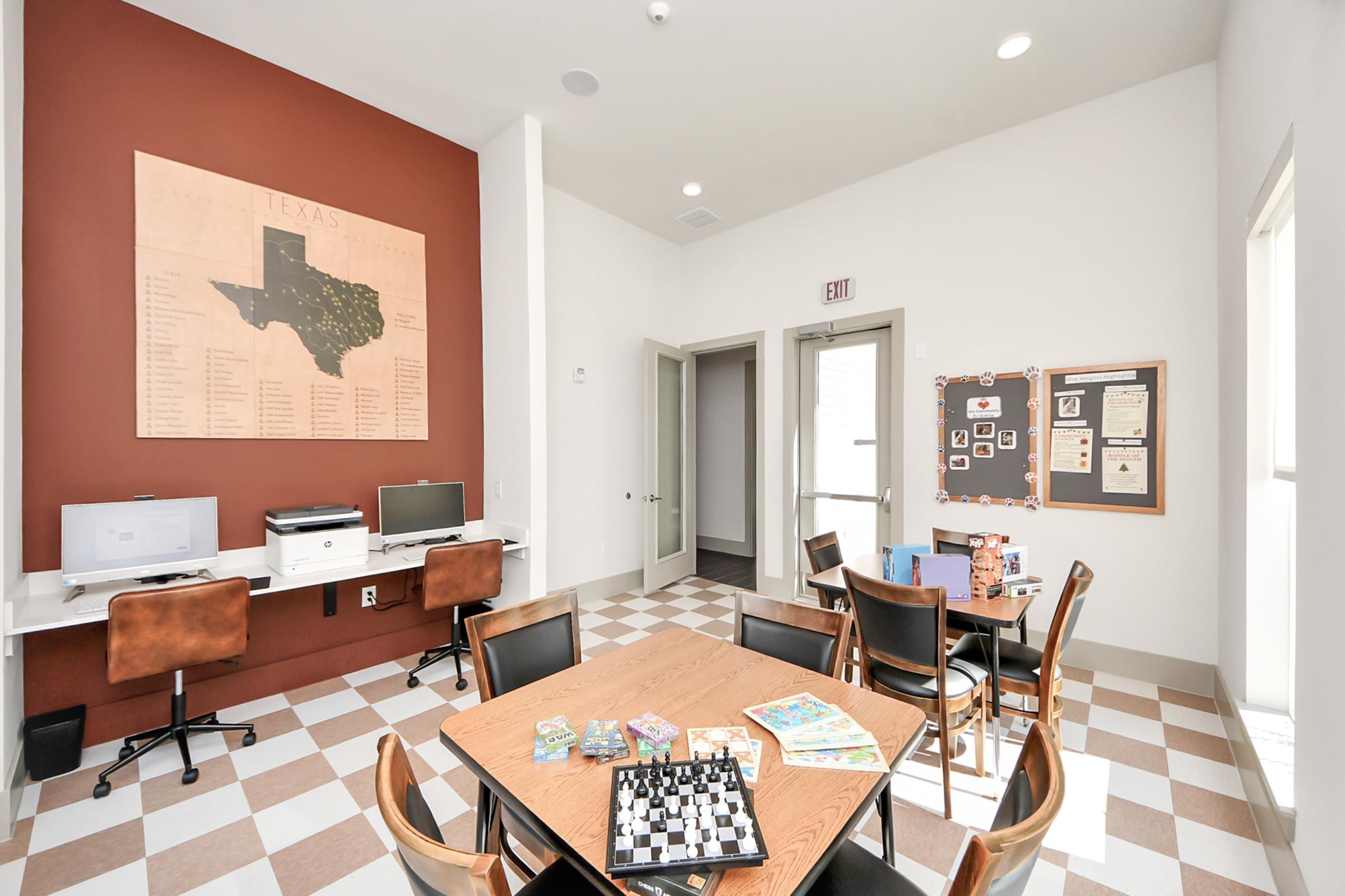 Interior of a room featuring a Texas map on the wall. There's a table with games and a chessboard in the center, surrounded by chairs. Two computers are positioned on a desk against one wall. The space has a modern design with a mix of light and dark colors. Bright lighting creates an inviting atmosphere.
