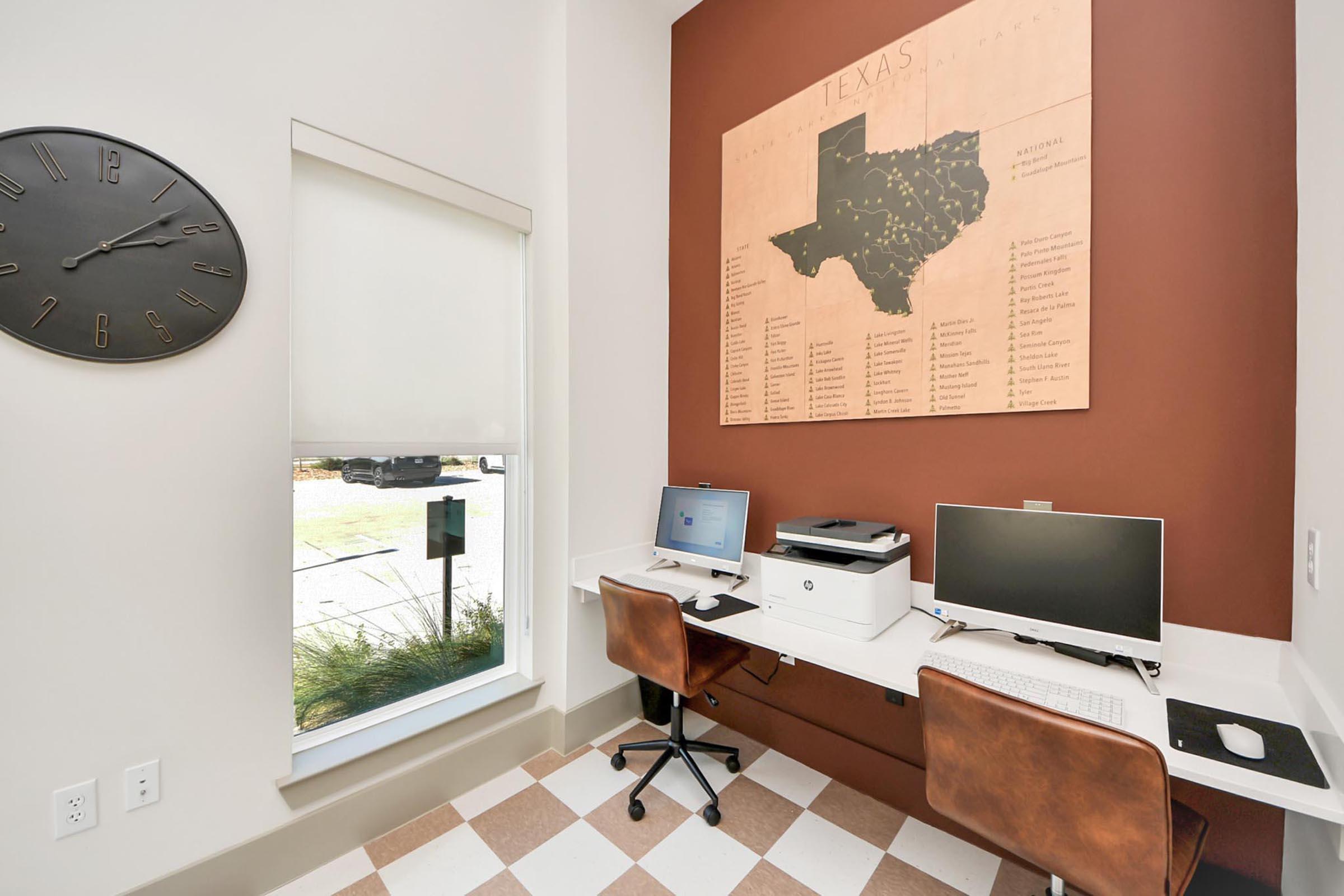 A small office nook featuring two computer workstations with brown leather chairs, a printer, and a wall-mounted map of Texas. There's a large clock on the wall and a window with a roller blind, allowing natural light into the space. The floor is checkered with light and dark tiles.