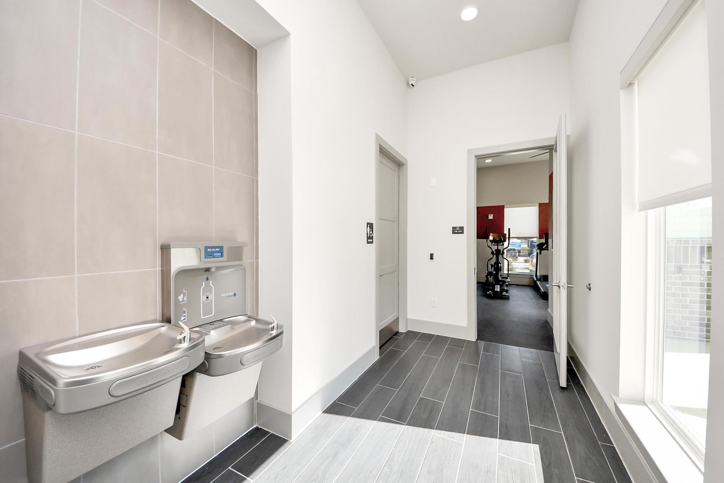 Interior view of a modern facility showcasing two drinking fountains on the left against a tiled wall. A doorway on the right leads to a fitness area, partially visible with exercise equipment. The space is bright, featuring large windows and a clean design with gray flooring.