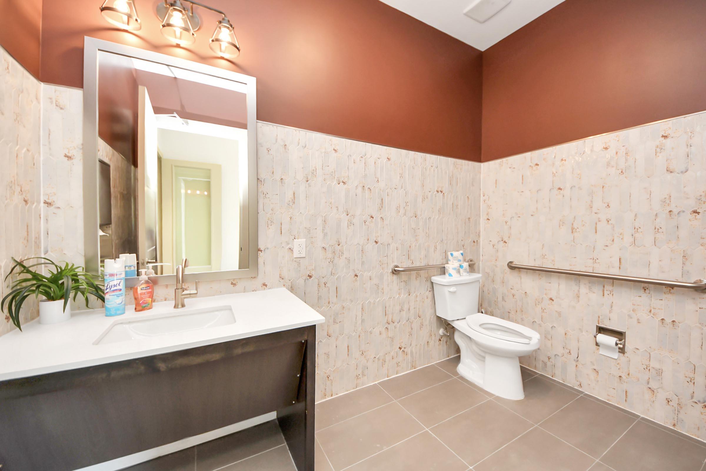 A modern restroom featuring a white toilet, a large mirror with decorative lighting, a stylish sink with a dark wooden vanity, and light-colored walls combined with dark accents. The space is clean and well-lit, with some toiletries visible on the sink and a small plant on the counter.