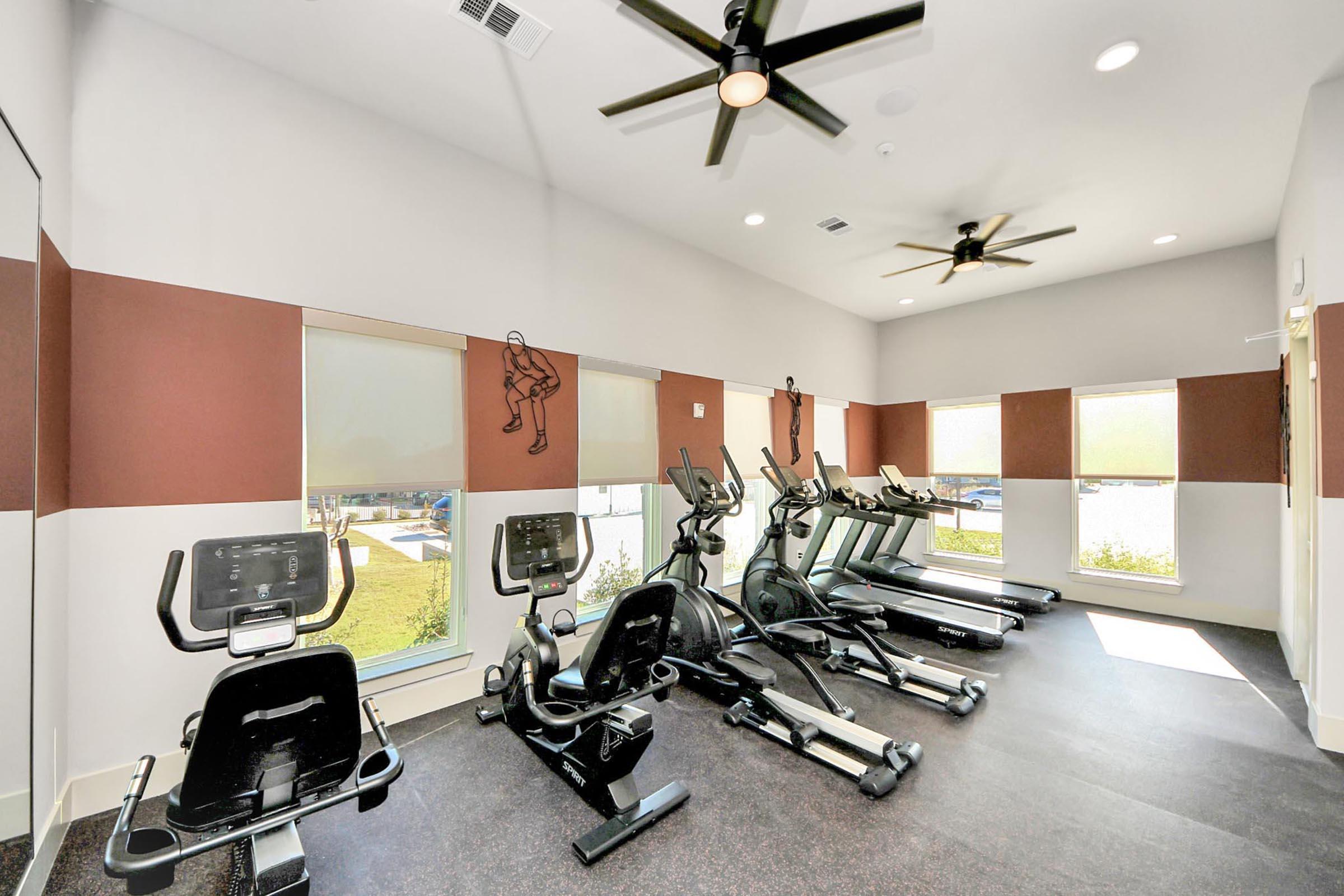 A modern gym interior featuring several elliptical machines and stationary bikes, with large windows allowing natural light. The walls are painted in a light color with maroon accents, and there are ceiling fans providing ventilation. The floor is covered with dark flooring for a sleek appearance.