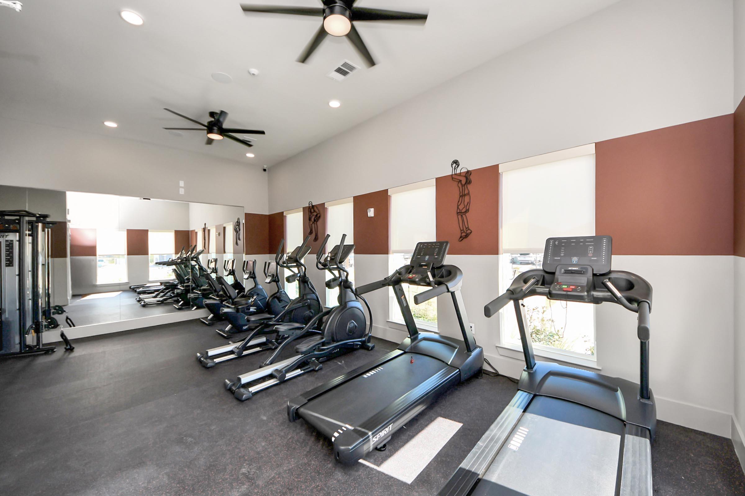 A modern gym interior featuring several treadmills lined up against the wall, large windows allowing natural light, a wall-mounted mirror for reflection, and ceiling fans for ventilation. The color scheme includes neutral walls with accents of dark brown.
