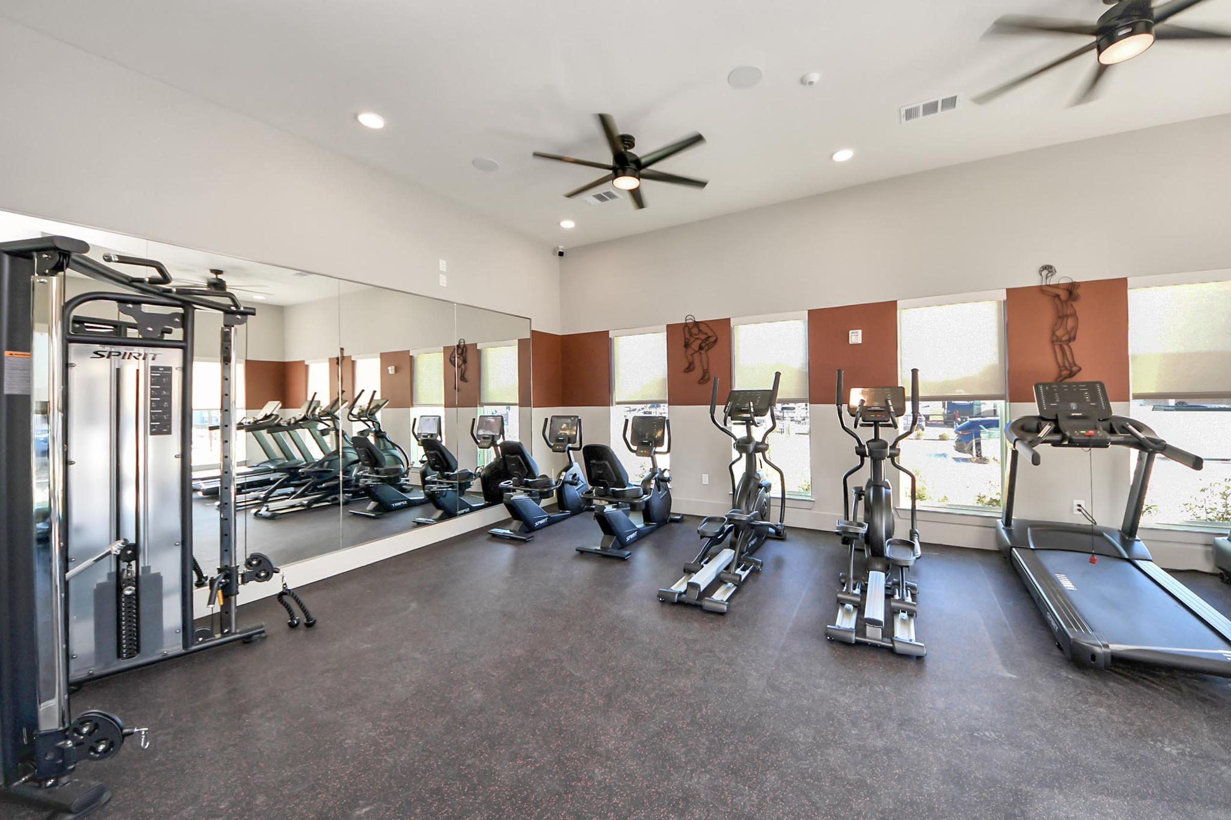 A modern gym interior featuring several exercise machines, including treadmills and ellipticals. Large windows provide natural light, and there are mirrors on one wall. The flooring is dark, and the space has ceiling fans for ventilation, creating a bright and inviting atmosphere for workouts.