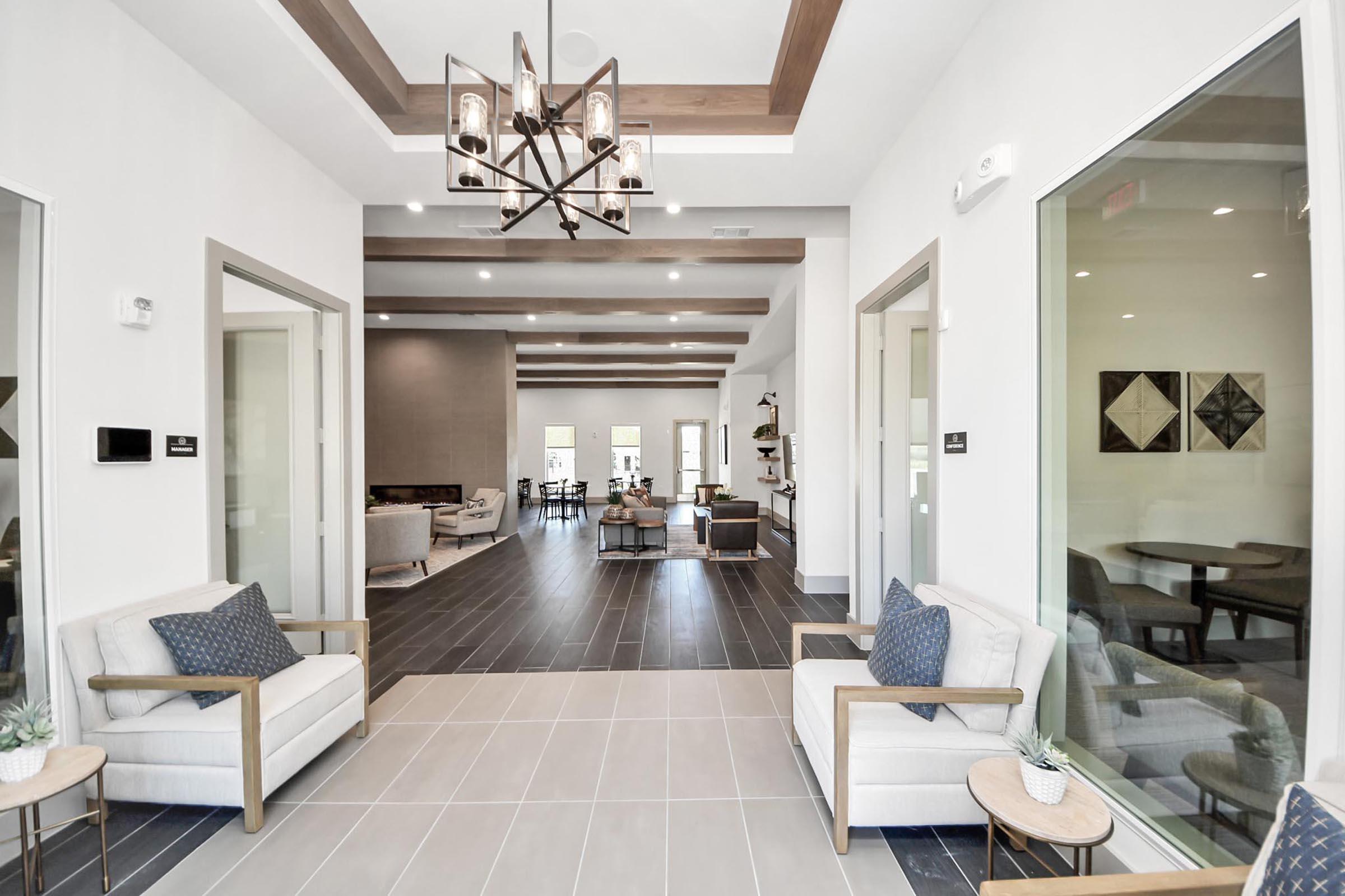A modern interior space featuring a welcoming entrance with two light-colored armchairs and decorative pillows. The room is well-lit, showcasing a stylish chandelier and a view into a spacious lounge area with contemporary furniture and art pieces, framed by clean lines and neutral tones.