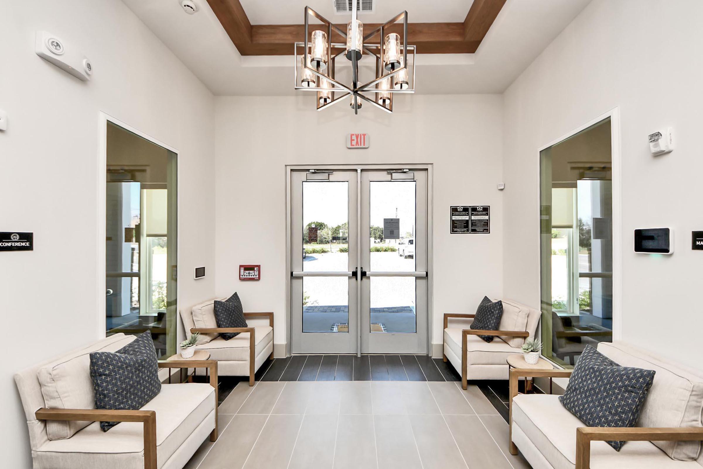 A modern lobby featuring two beige armchairs with decorative pillows, a stylish chandelier, and large glass doors leading outside. The walls are painted white, and there are small plants on the chairs. Signs and a window are also visible, adding to the inviting atmosphere.
