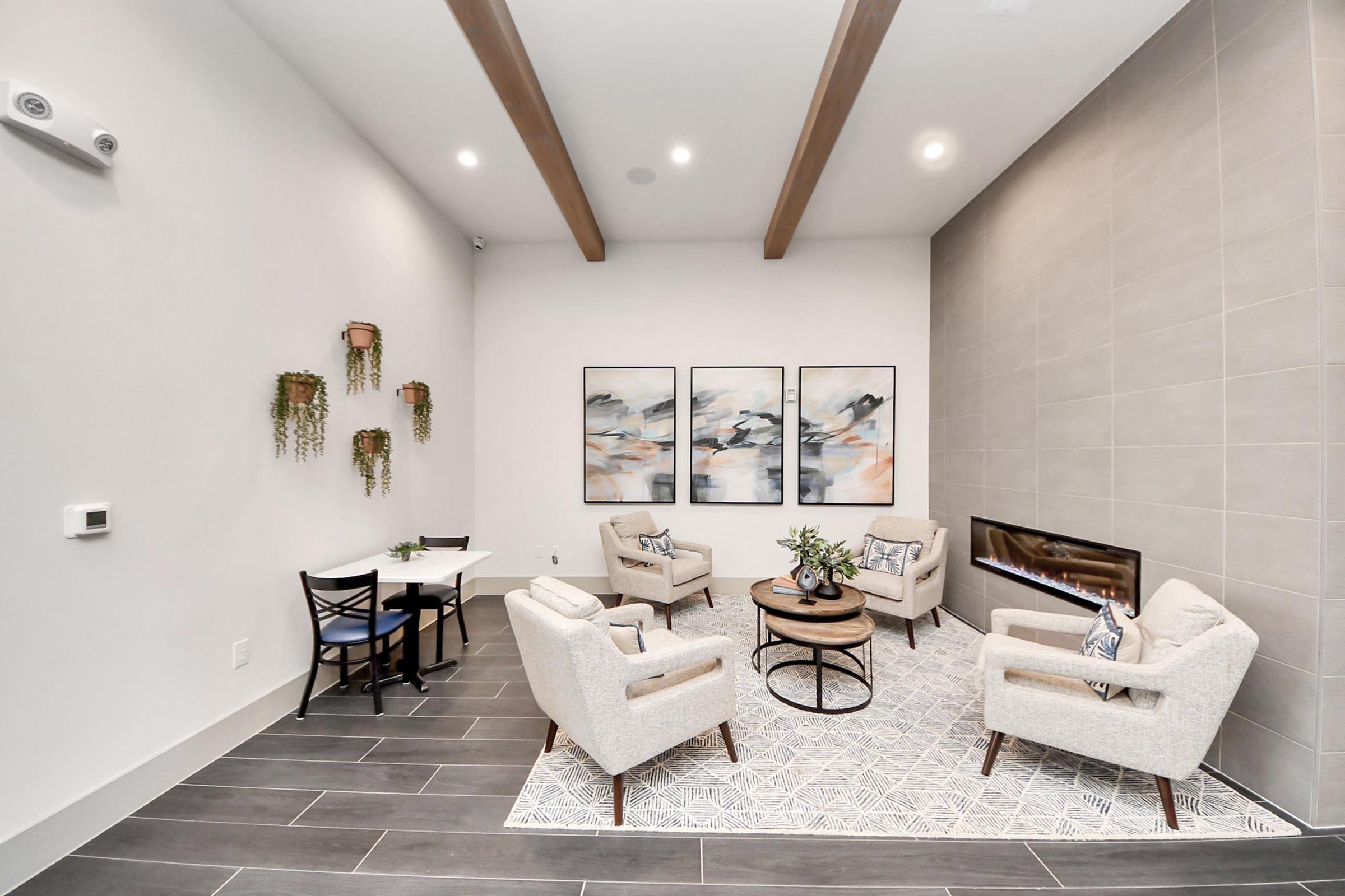 A modern living space featuring light-colored walls and dark tiled flooring. It includes two stylish armchairs and a round coffee table on a patterned rug. There is a small dining table for two, wall-mounted plants, and three abstract wall art pieces. A sleek fireplace adds warmth to the room.