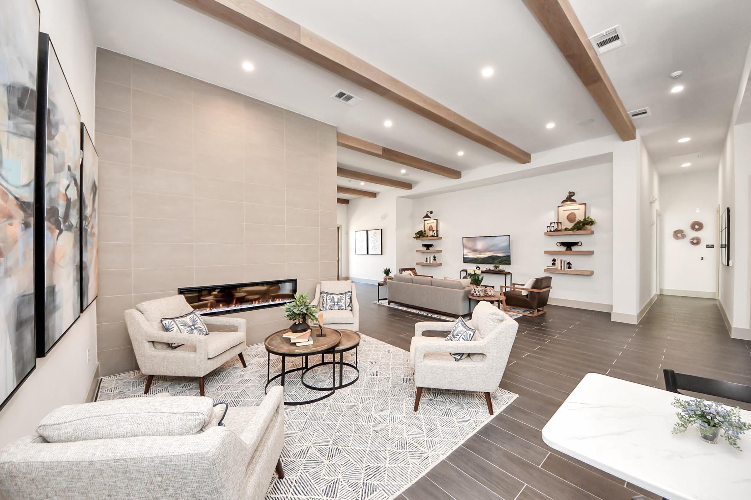 Spacious modern living room featuring light-colored walls and a large window. Two comfortable chairs and a round coffee table are positioned on a patterned rug. A sleek fireplace is centered on the wall, with decorative shelves and a TV nearby. The ceiling has wooden beams for added style and warmth.