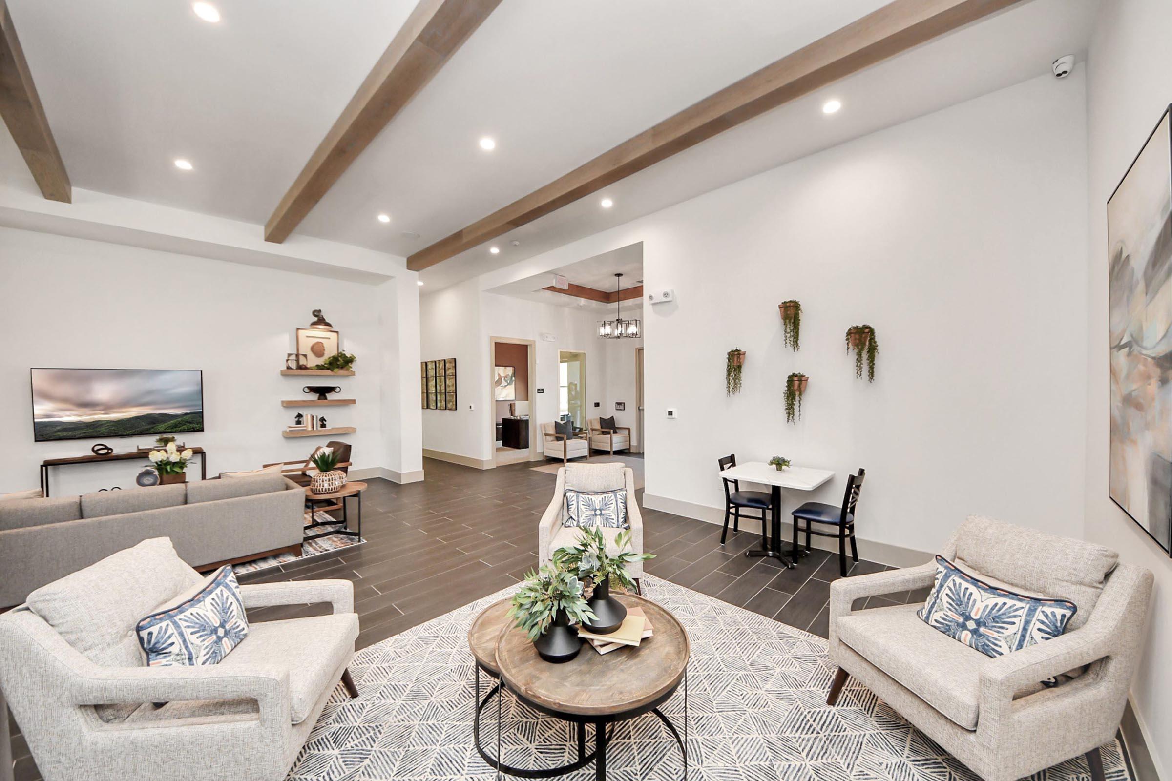 Modern living room with a cozy seating arrangement featuring two light-colored armchairs and a circular coffee table. A flat-screen TV on the wall and decorative plants add to the ambiance. The space has light walls, wooden beams on the ceiling, and a small dining area visible in the background.