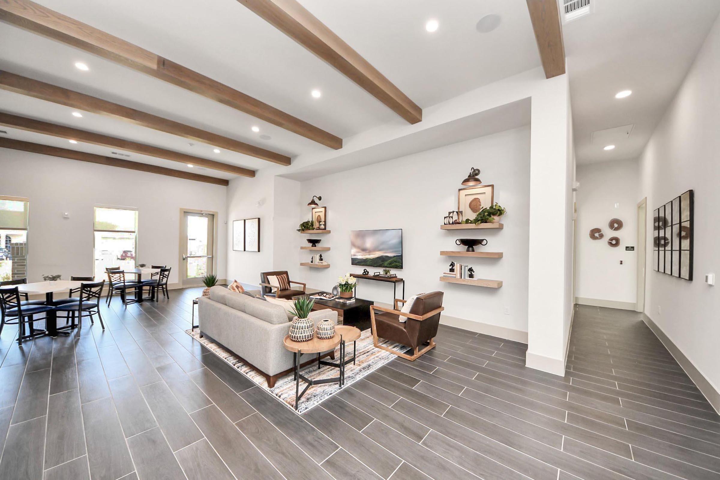 A modern living space featuring a cozy seating arrangement with a light gray sofa, two dark chairs, and wooden shelves displaying decorative items. The room has wooden beams on the ceiling, large windows allowing natural light, and a stylish rug, creating a warm and inviting atmosphere.