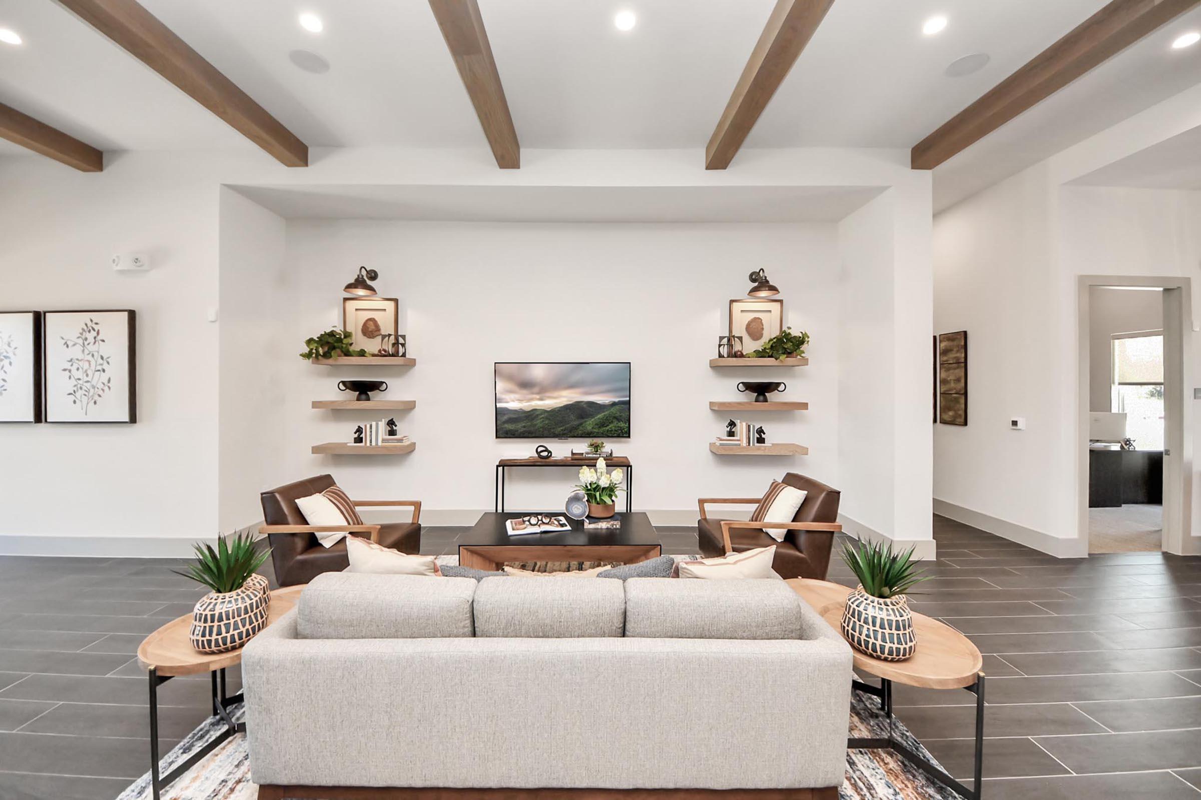Modern living room featuring a light-colored sofa with decorative pillows, two stylish armchairs, and a wooden coffee table. Wall-mounted shelves display decorative items and a flat-screen TV is centered on the wall. The room has neutral colors and wooden beams on the ceiling, creating a spacious and inviting atmosphere.