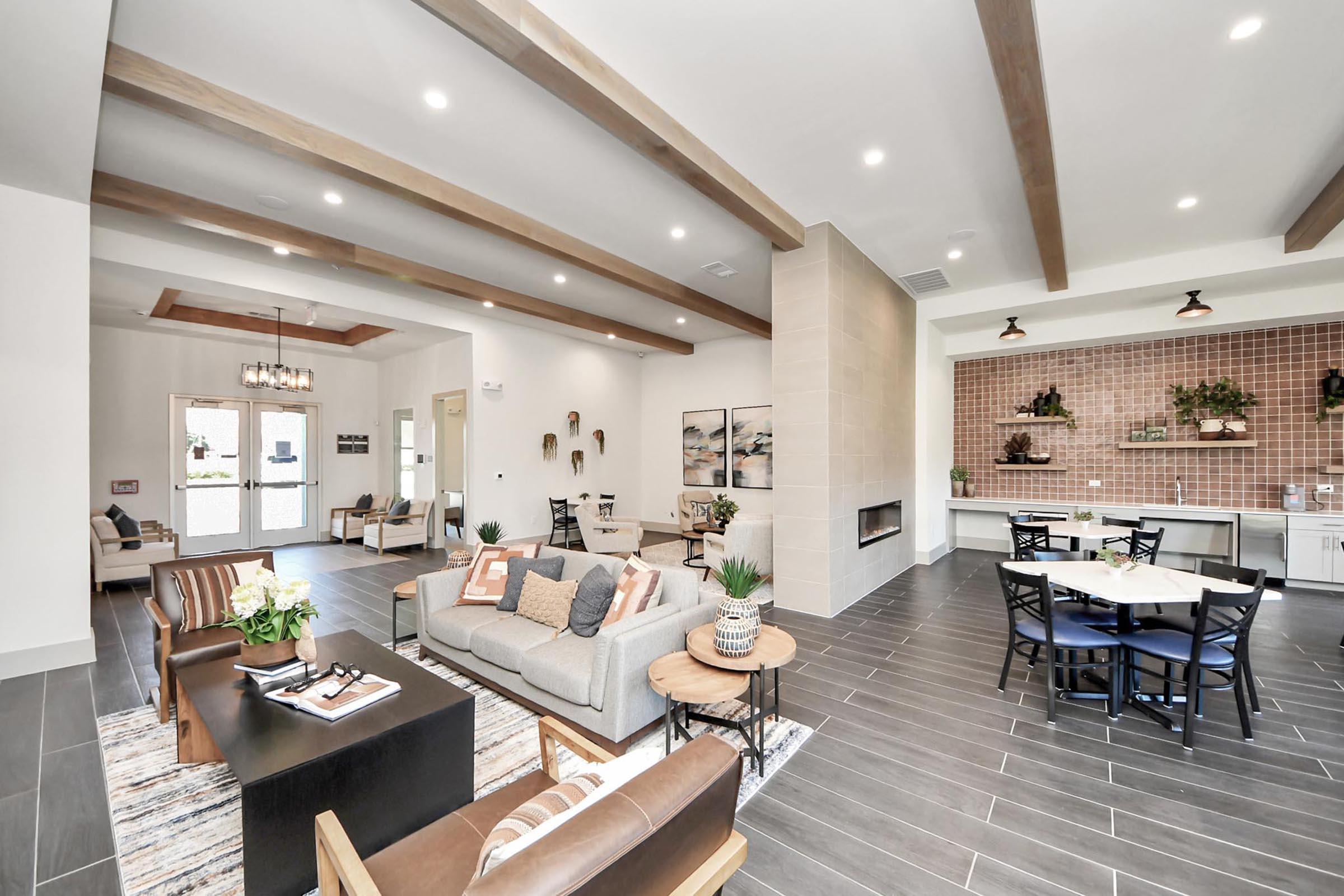 A spacious modern living area featuring stylish furniture, including a gray sofa and a wooden coffee table. There are decorative plants and artwork on the walls, with a cozy dining space in the background. The space has high ceilings, exposed wooden beams, and large windows allowing natural light to fill the room.