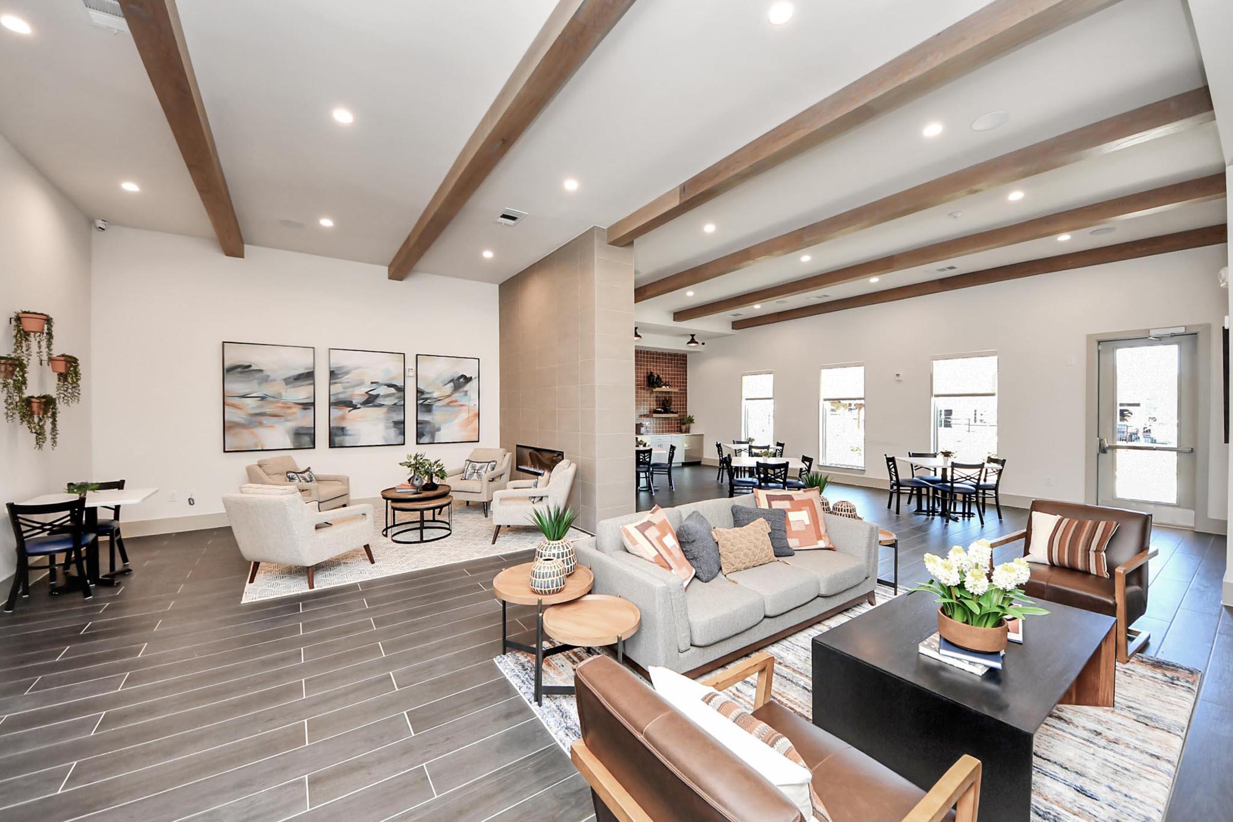 A modern, spacious common area featuring comfortable seating with neutral-toned furniture, decorative plants, and artwork on the walls. Natural light fills the room through large windows, highlighting the wooden beams on the ceiling. There are dining tables and chairs in the background, creating a welcoming atmosphere.