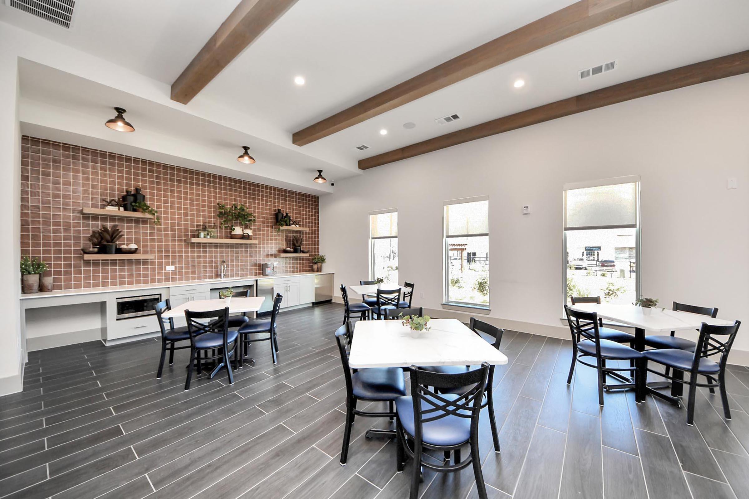 Modern dining area with four tables, each set with small plants. Features a stylish wall with brown tile and decorative plants. Large windows let in natural light, and the space has wooden beams overhead, creating a warm and inviting atmosphere.