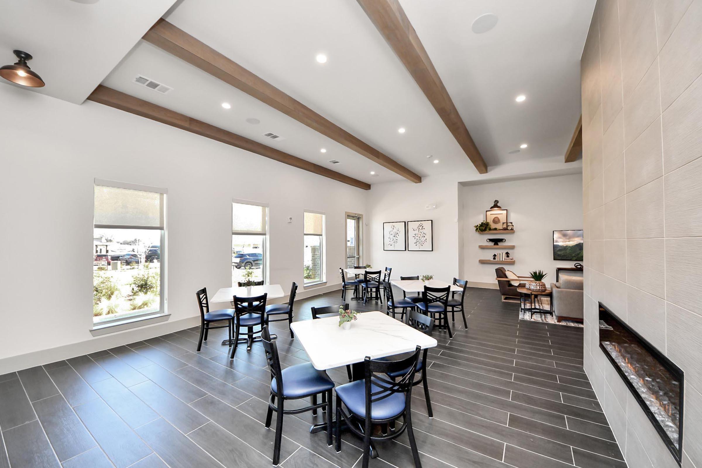 A modern, well-lit interior of a common area featuring a white marble table with chairs, a decorative fireplace, and large windows. The wall has wooden beams and minimalistic decor, creating a welcoming and stylish atmosphere. There are plants visible outside the windows.