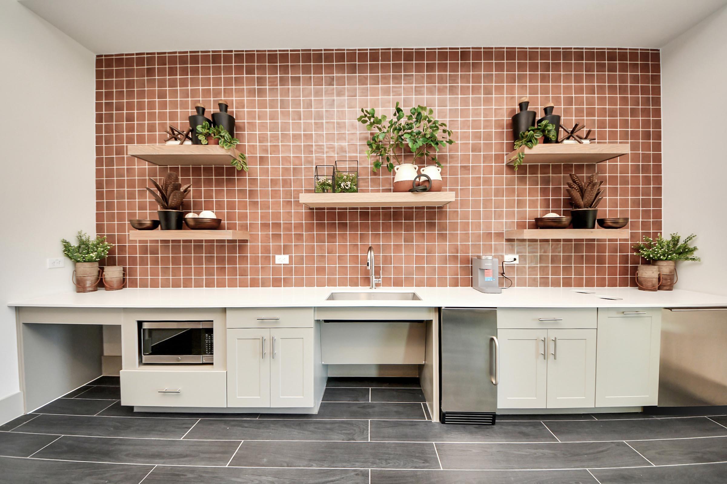 A modern kitchen featuring a pink tiled backsplash, white countertops, and open shelving adorned with potted plants. The space includes a stainless steel sink, a microwave built into the cabinetry, and dark tile flooring, creating a stylish and inviting atmosphere.