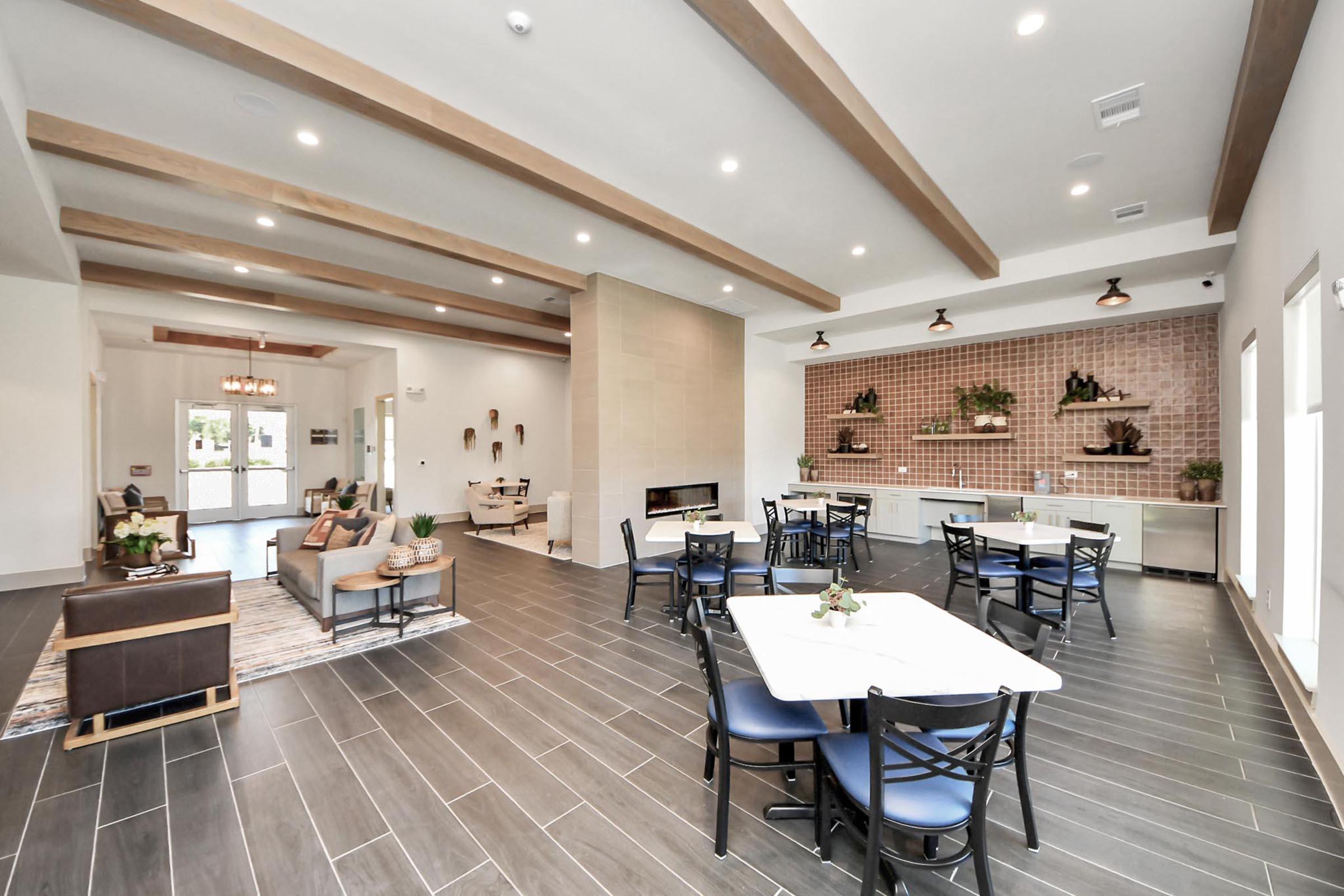 A modern interior of a spacious room featuring wooden beams, a cozy seating area with a couch and coffee table, and tables with chairs for dining. There is a fireplace, decorative wall shelves, and stylish lighting fixtures, creating a welcoming atmosphere.