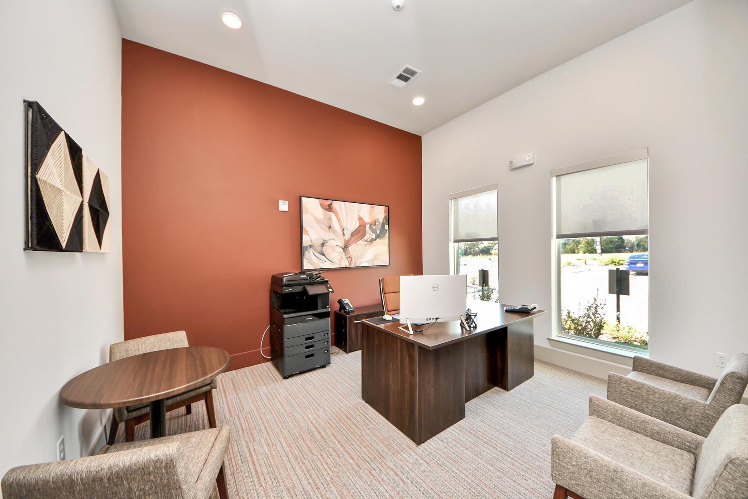 A modern office space featuring a brown accent wall, a desk with a computer, a printer, a telephone, and a small round table with two chairs. The room is well-lit with large windows and has a minimalist decor, including abstract wall art and a light-colored carpet.