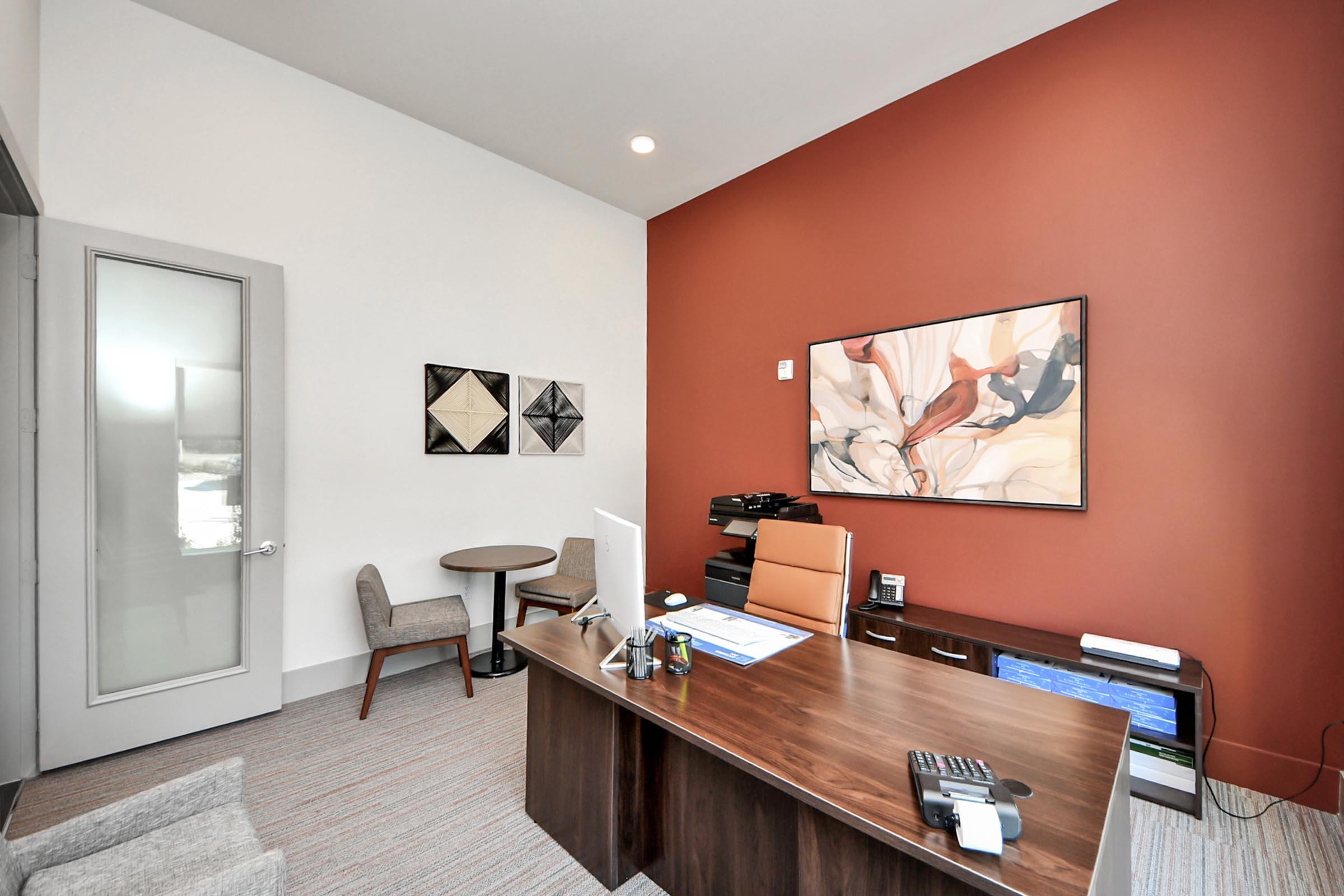 Modern office interior featuring a wooden desk with a computer, an orange office chair, and a phone. There are two gray upholstered chairs and a small round table in the corner. The walls are a mix of white and a reddish-brown accent, with contemporary artwork hung above the desk. Natural light illuminates the space.