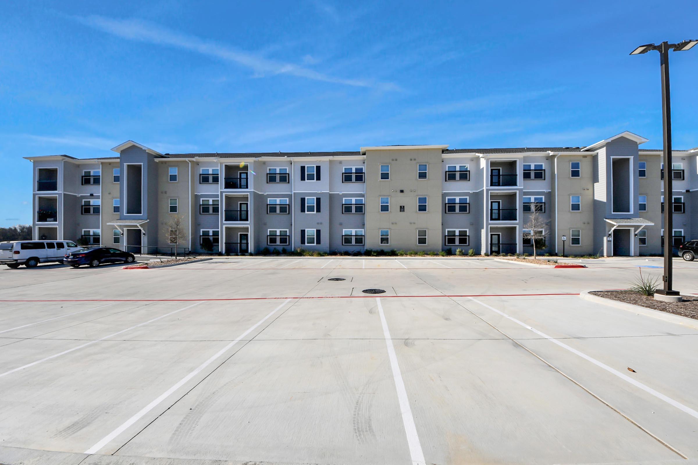 A modern apartment complex featuring multiple stories, with a light gray and white exterior. The building includes balconies and large windows. In the foreground, a paved parking lot with several empty spaces is visible, under a clear blue sky.