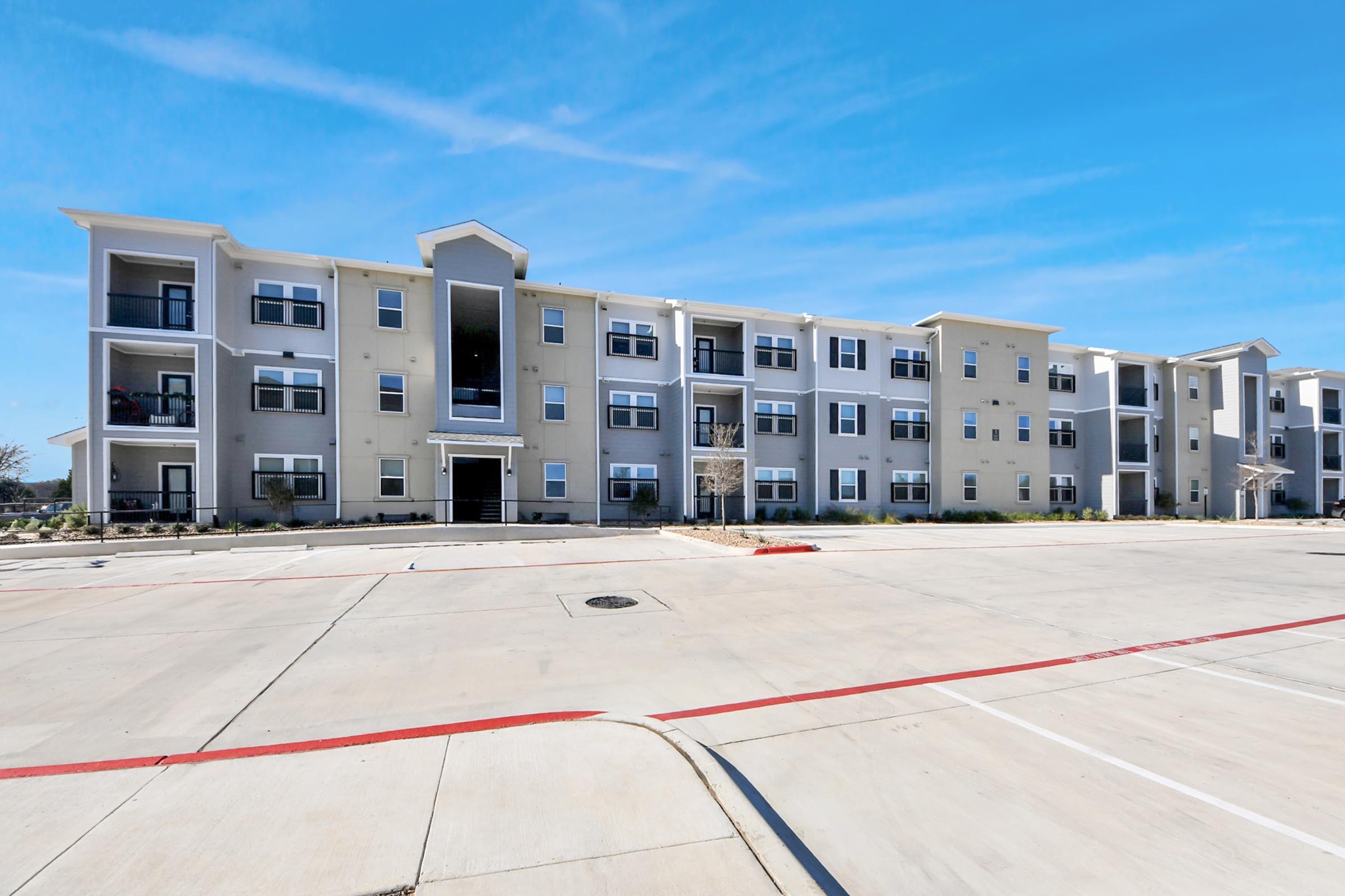 A modern three-story apartment building with a light tan exterior and large windows, featuring a central entrance. There is a spacious paved parking lot in front, with a red-painted curb. The sky is clear and blue, indicating a sunny day.