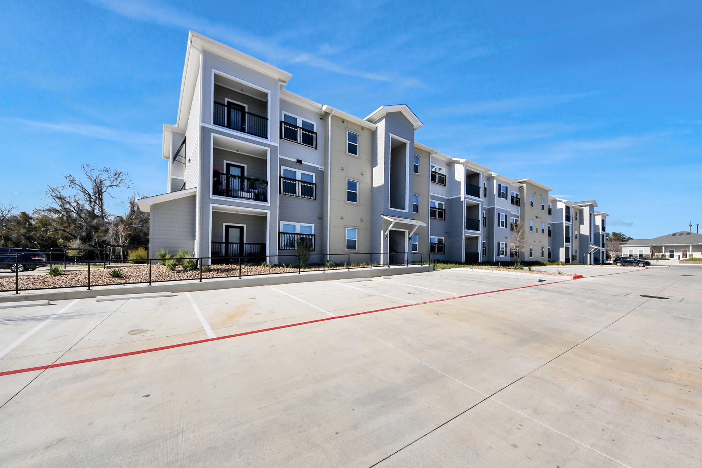 A modern multi-unit apartment building featuring a light-colored exterior, balconies, and a landscaped area. In front, there is a large parking lot with several empty spaces and clear blue skies overhead. The setting appears new and well-maintained.
