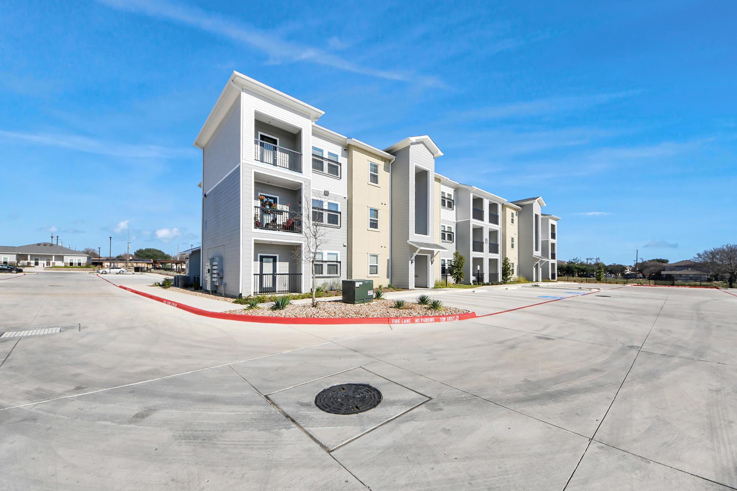 A modern multi-unit residential building with multiple stories and balconies, situated on a paved lot. The façade features a mix of light-colored materials, surrounded by landscaped areas. Clear blue sky overhead and a few parked vehicles are visible in the foreground.