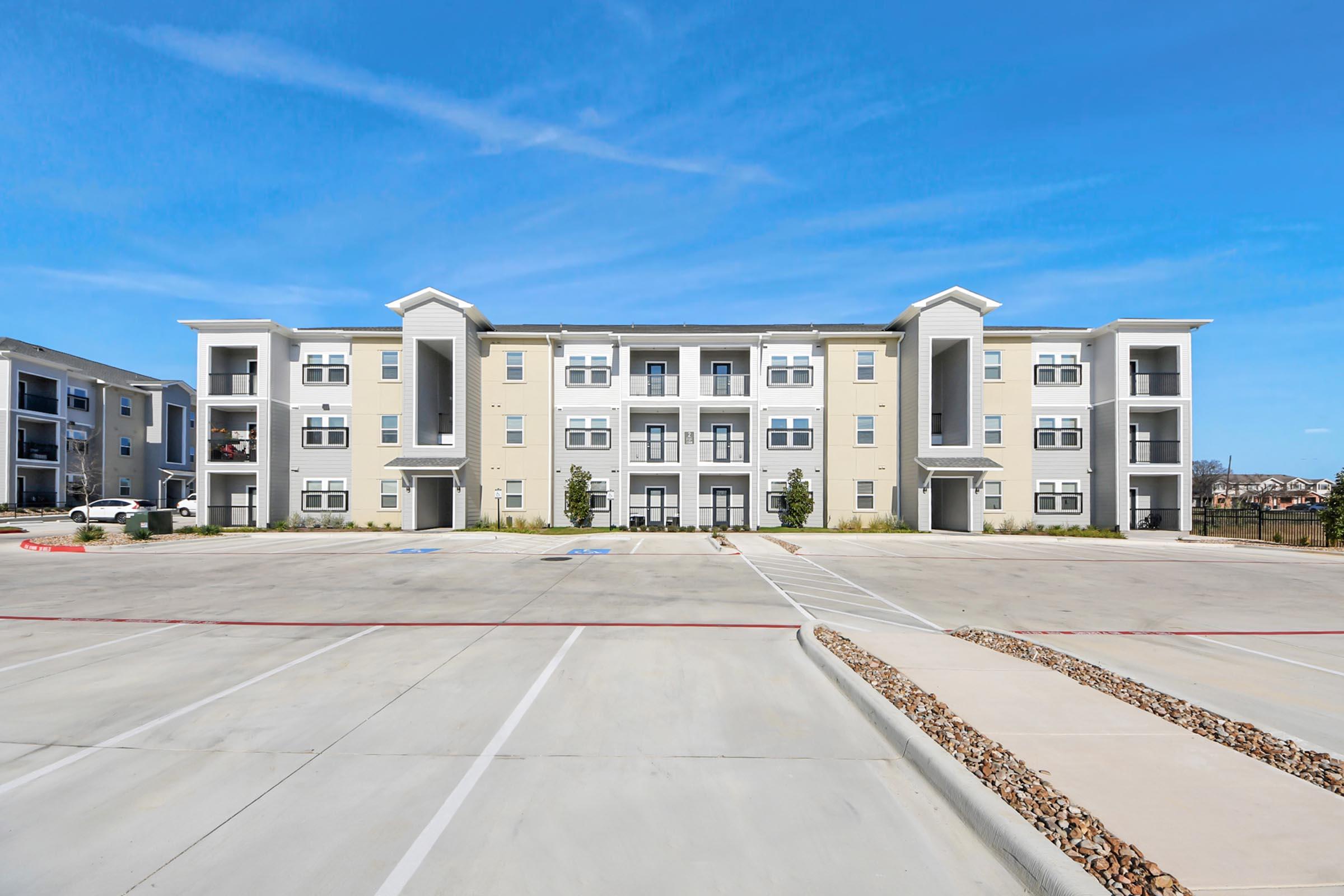 A modern, multi-story apartment building with a light exterior, featuring multiple balconies and large windows. The surrounding parking lot is clean and paved, with some landscaping and a clear blue sky above.
