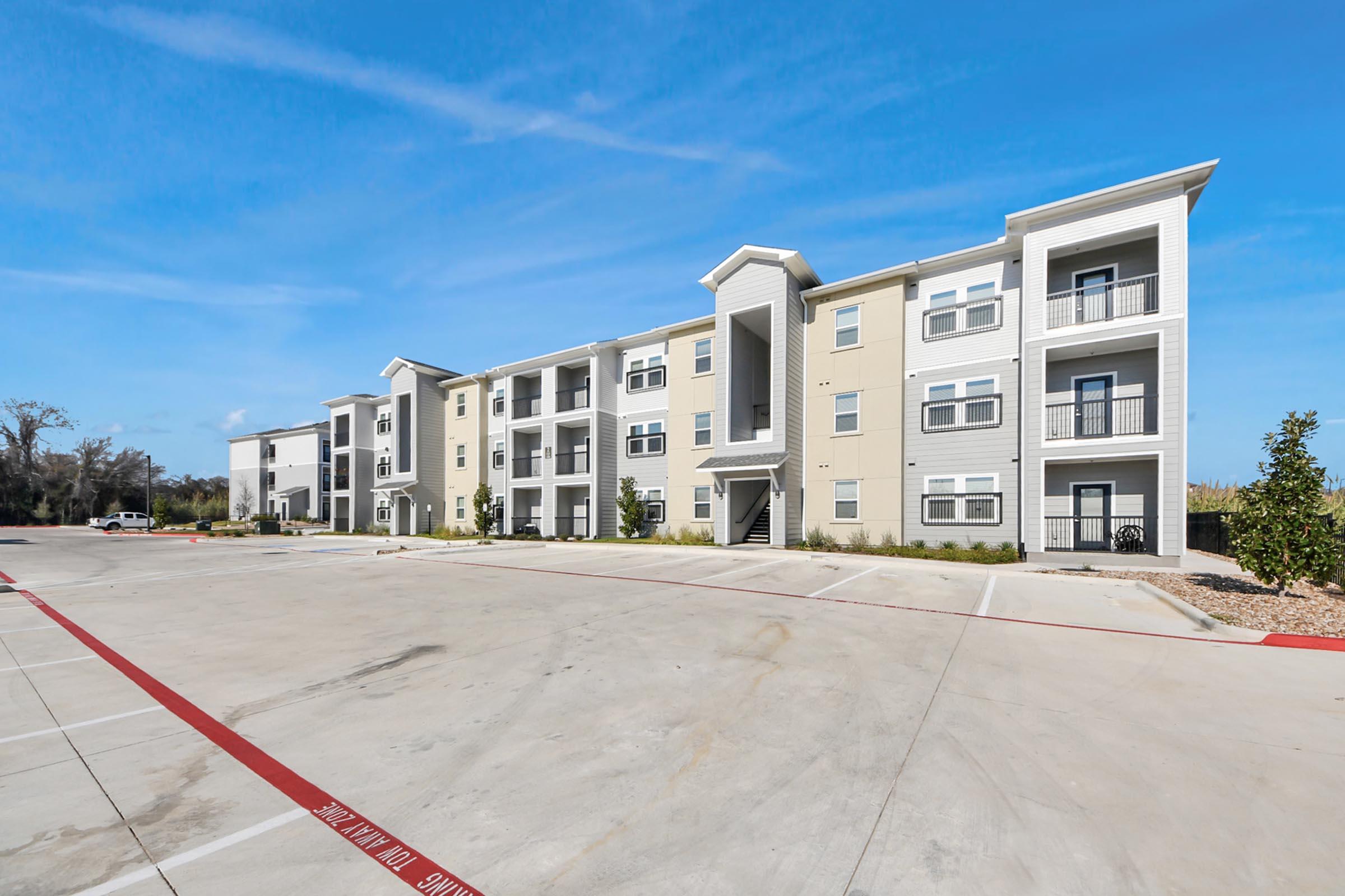 A modern three-story apartment building with beige siding, multiple balconies, and large windows, set against a clear blue sky. The surrounding area features a spacious paved parking lot with red striping, landscaping with small trees, and open space in the background.