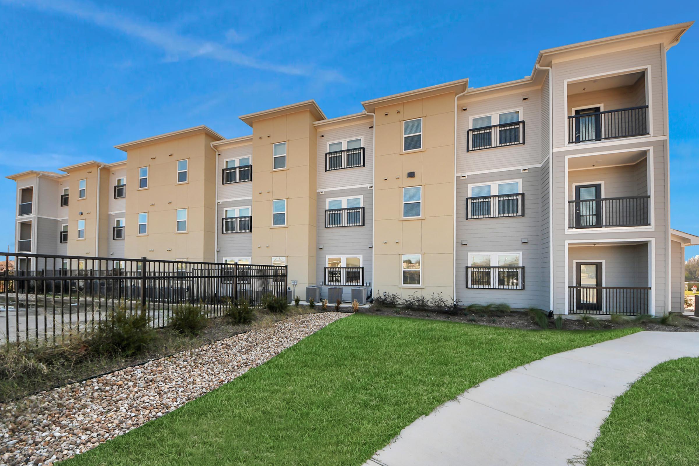 A modern multi-story apartment building featuring a combination of beige and gray siding. The exterior includes multiple balconies, large windows, and a landscaped area with a pathway and decorative stones. A black fence borders the property, set against a clear blue sky.