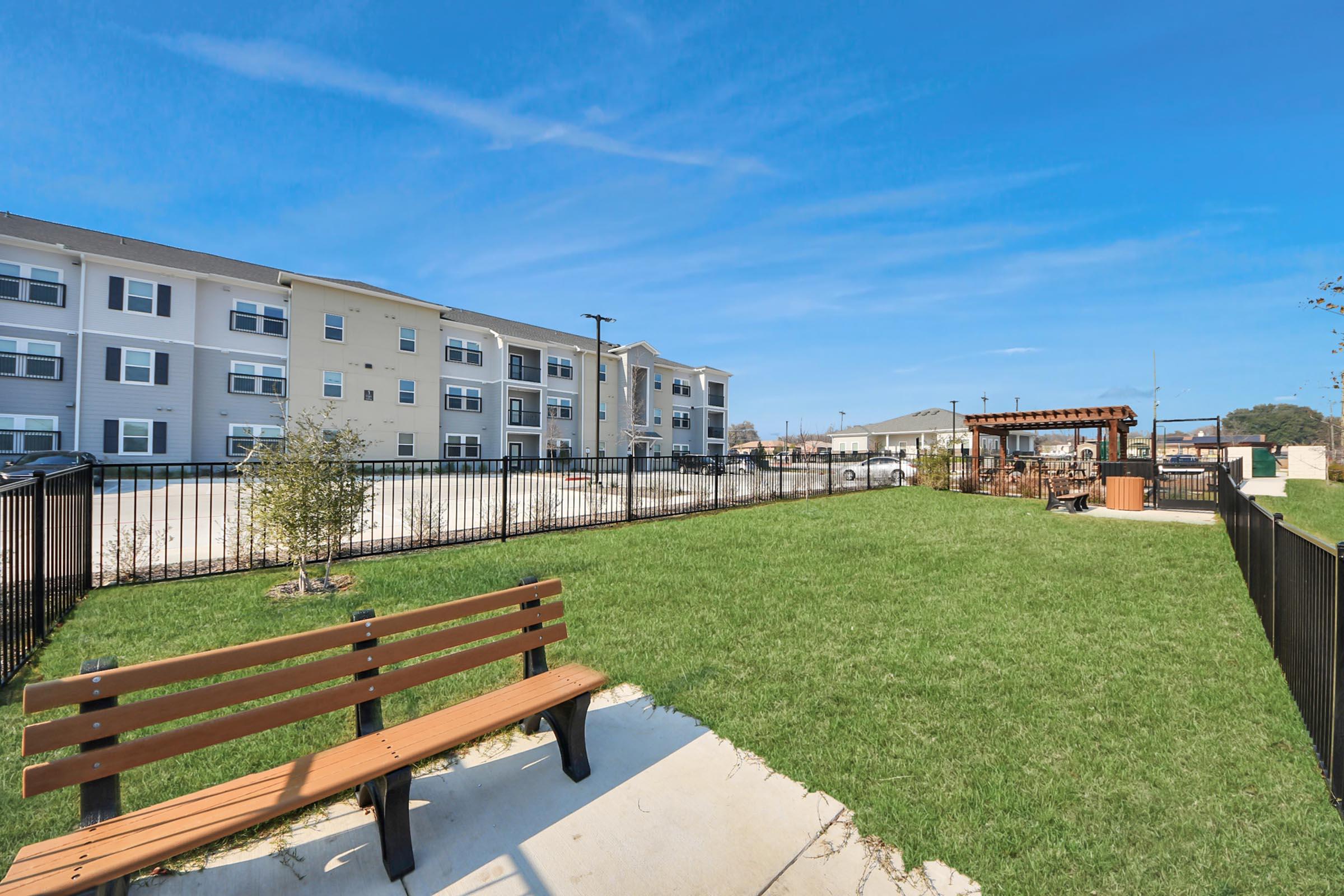 A spacious outdoor area featuring a green lawn, a wooden bench, and a fenced perimeter. In the background, there are modern apartment buildings and a patio area with seating. The sky is clear and blue, contributing to a bright and inviting atmosphere.