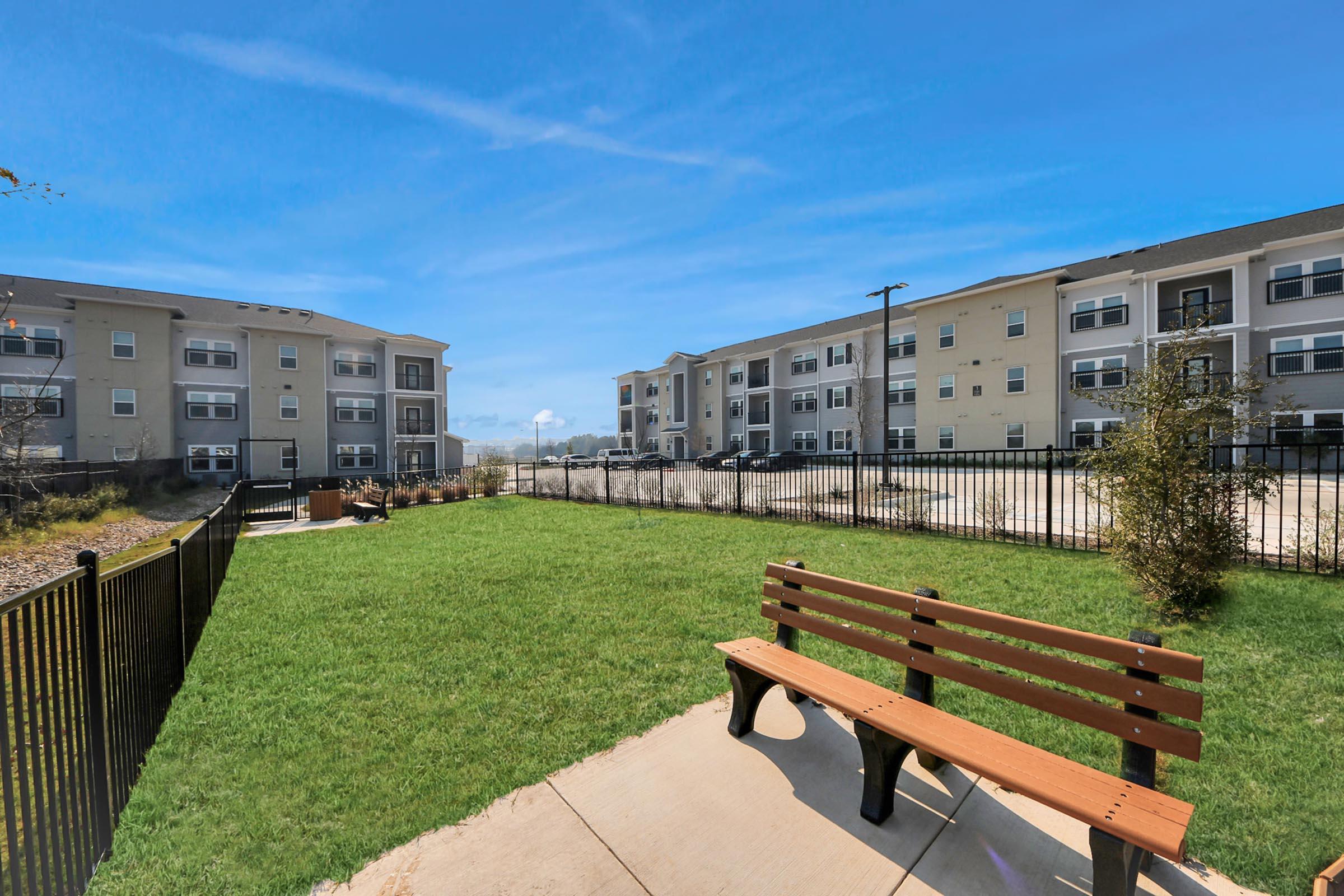 A green lawn with a wooden bench in the foreground, surrounded by a black fence. In the background, there are two multi-story apartment buildings with several parking spaces nearby and a clear blue sky overhead.