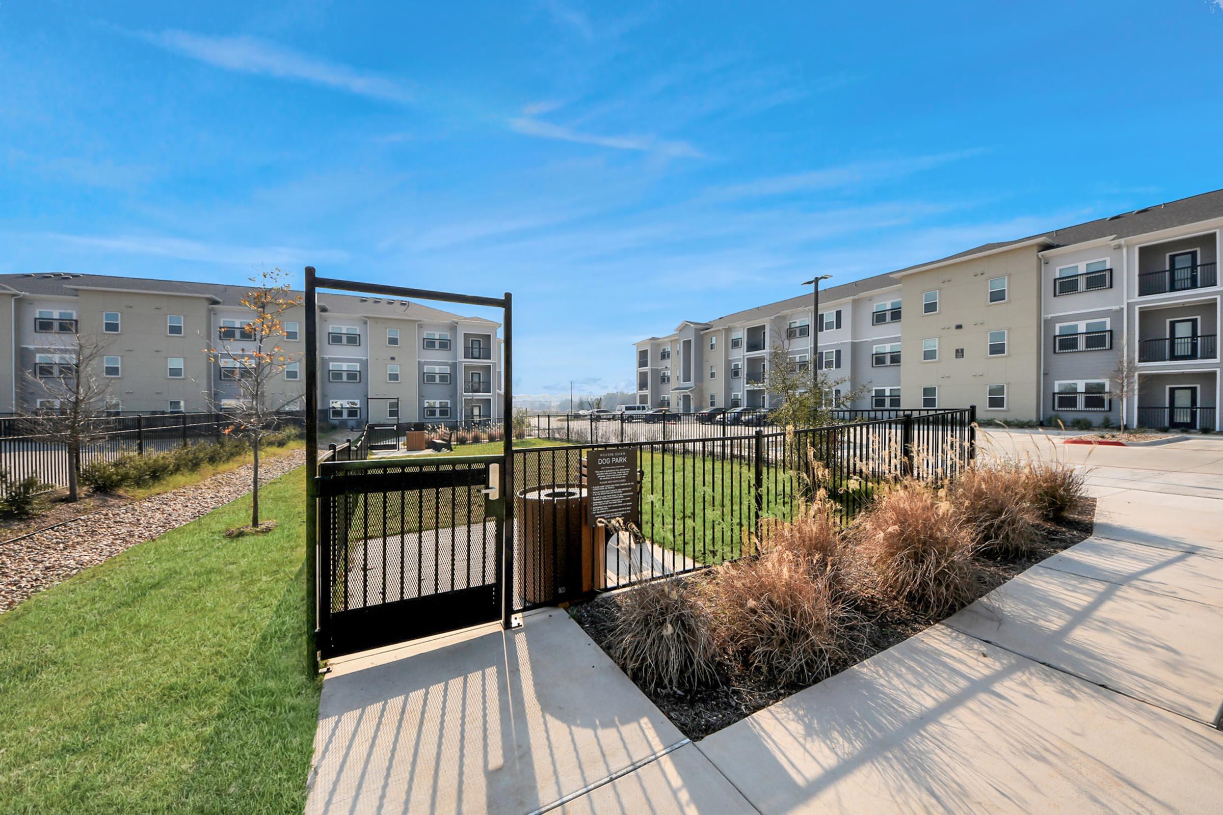 A gated entrance leading to a landscaped area with grass and bushes, surrounded by modern apartment buildings. The sky is clear and blue, enhancing the welcoming atmosphere of the residential complex.