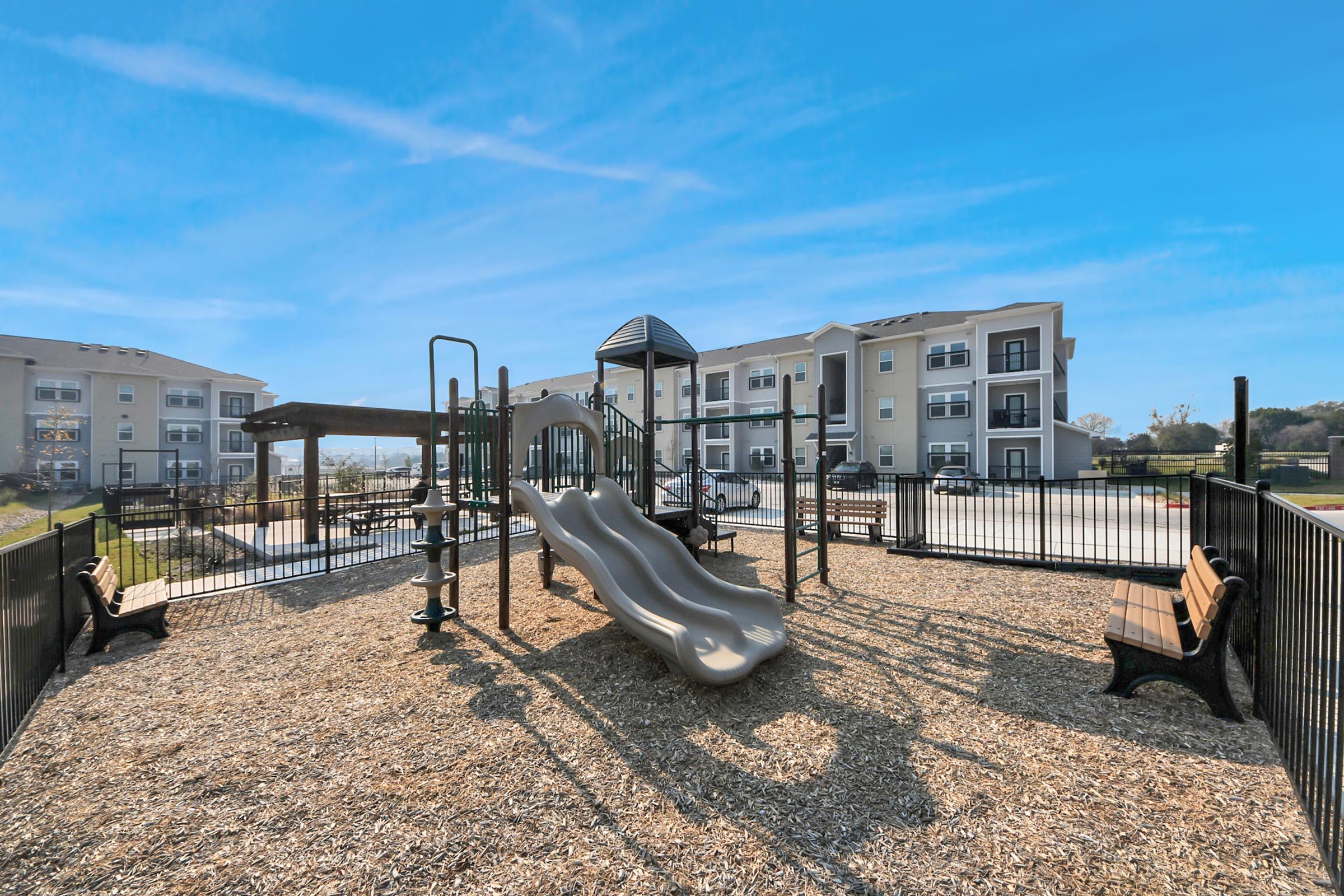A playground featuring two silver slides, climbing structures, and benches surrounded by a safety fence. In the background, there are two multi-story residential buildings and a clear blue sky. The ground is covered with wood chips for safety.