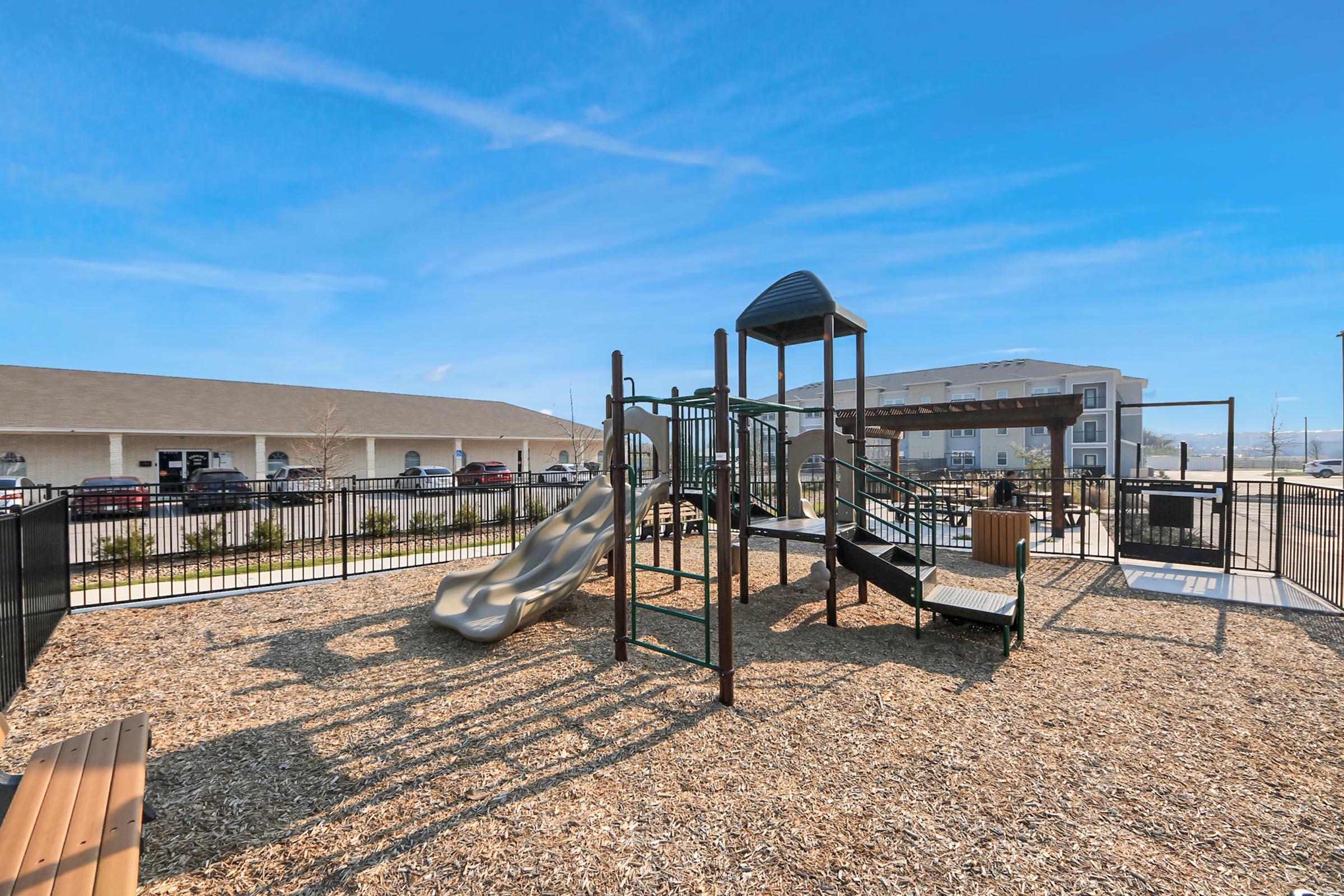 A playground featuring a multi-level structure with slides, climbing equipment, and a sandbox area, surrounded by safety landscaping. In the background, there is a building and parked cars under a clear blue sky, indicating a sunny day.