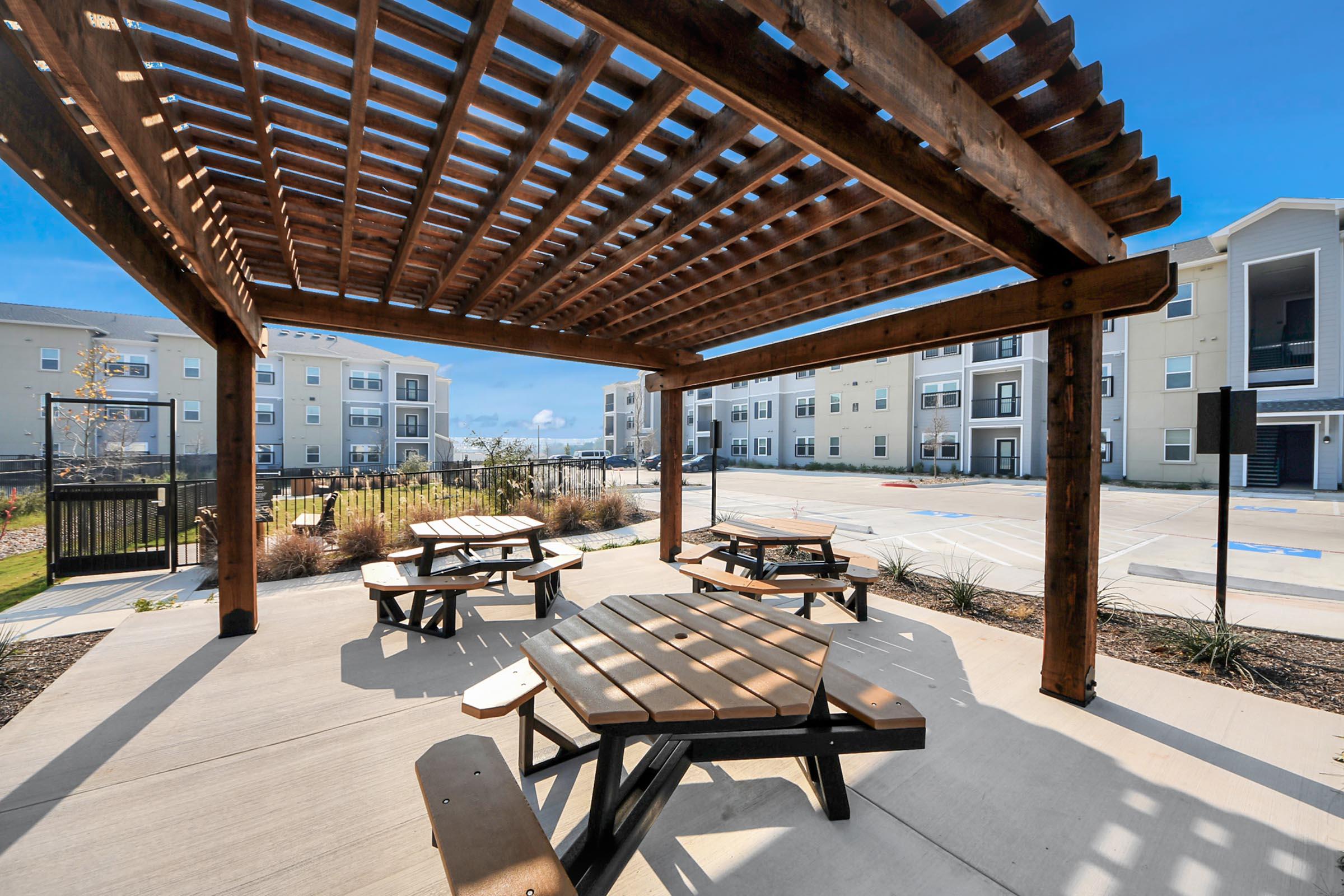 Outdoor seating area featuring wooden picnic tables under a pergola. The background shows modern apartment buildings and a clear blue sky, creating a welcoming ambiance for relaxation or social gatherings. Surrounding greenery adds to the inviting atmosphere.