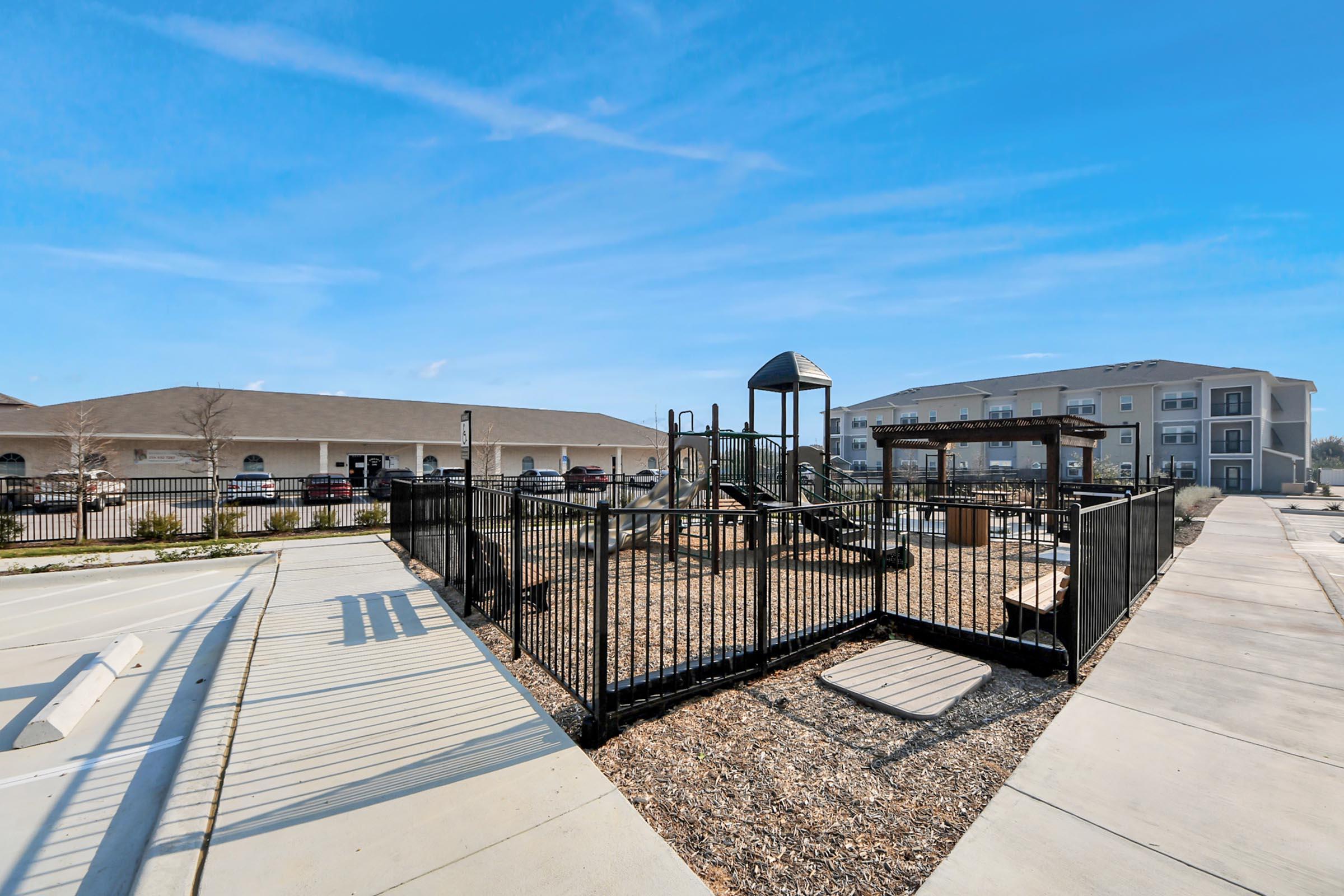 A fenced playground area featuring a climbing structure and a sandbox, surrounded by a paved walkway. In the background, there are residential buildings and parked cars under a clear blue sky. The scene is bright and inviting for outdoor play.