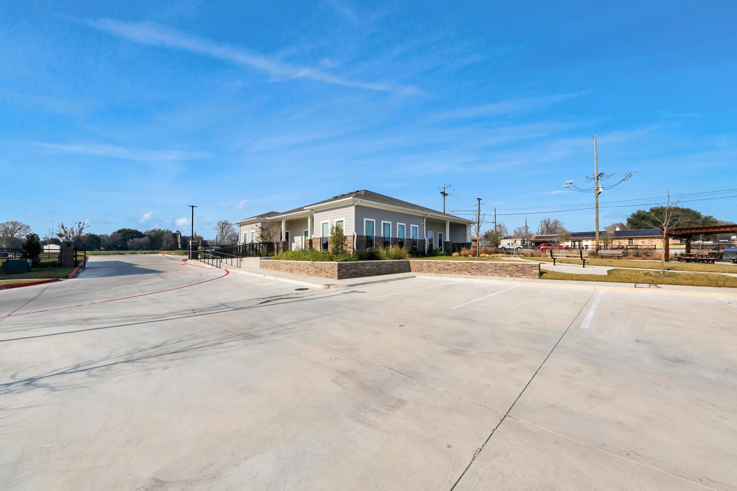 A modern building with a light-colored exterior sits on a wide, paved area. The scene features clear blue skies and a few scattered clouds, with an empty parking lot in the foreground. Surrounding greenery and utility poles are visible, creating a calm suburban atmosphere.