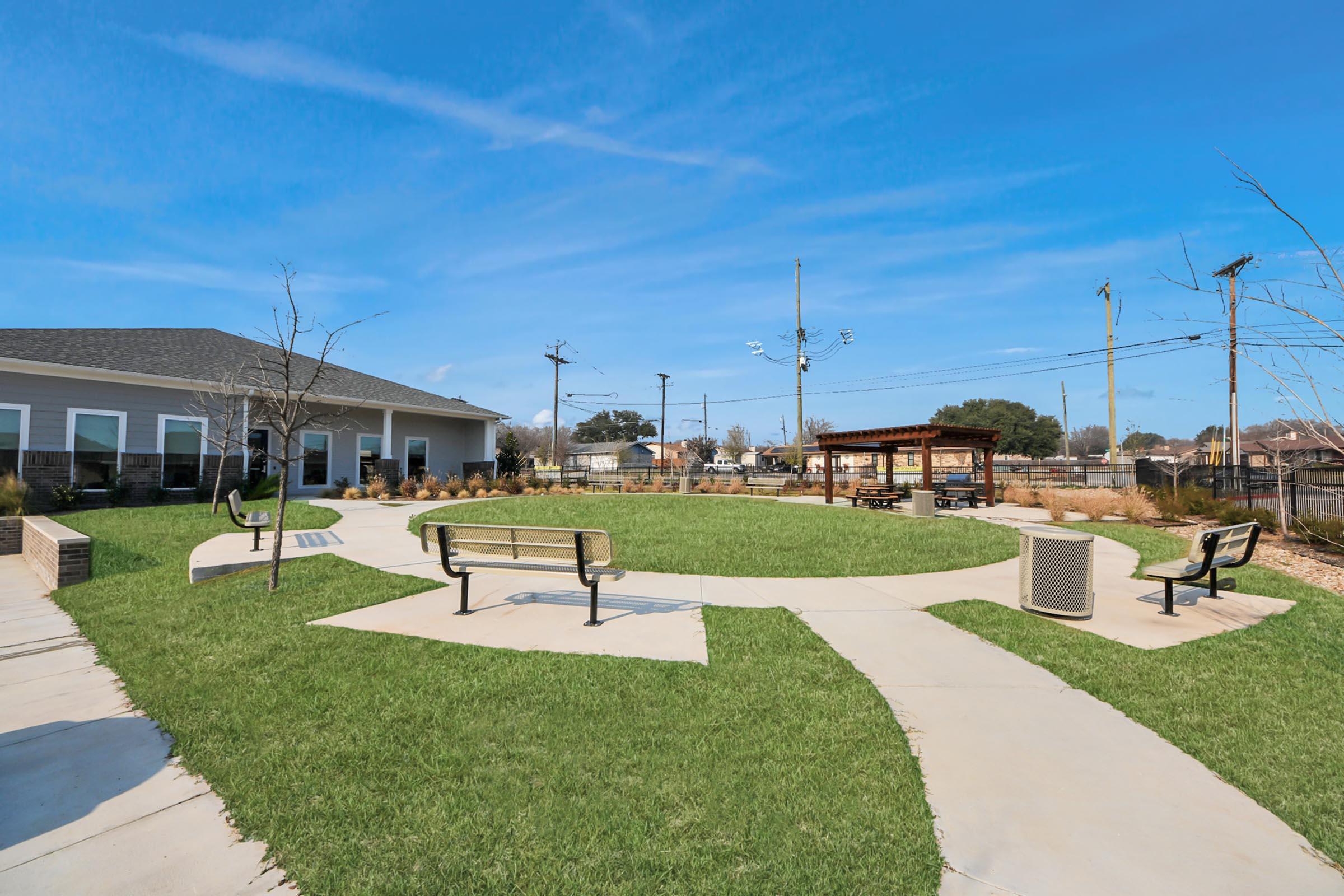 A well-maintained outdoor area featuring green grass, curved pathways, and benches. In the background, there is a small gazebo and a building with a gray roof. The sky is clear and blue, and there are utility poles in the distance. The space is designed for relaxation and social gatherings.