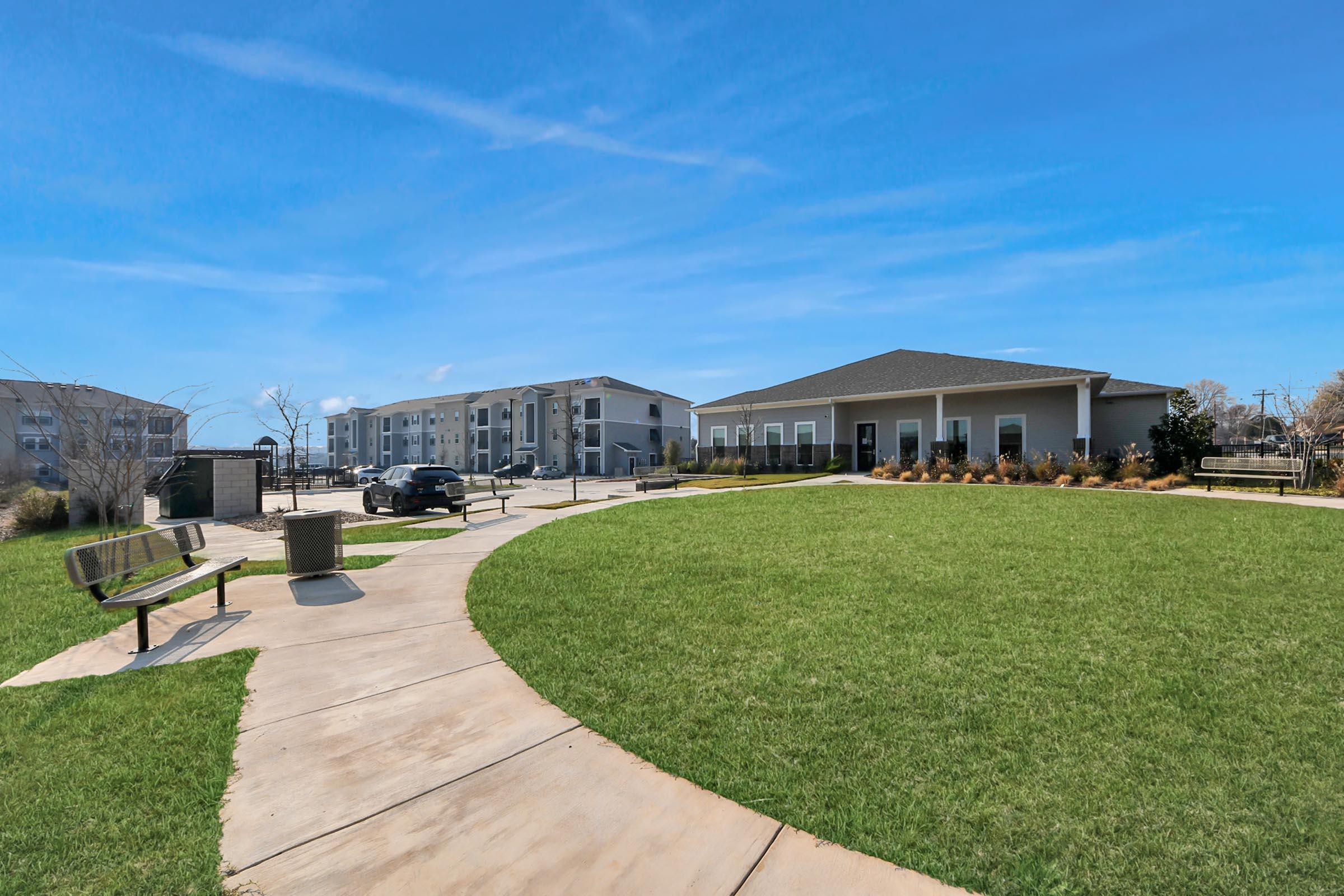 A well-maintained park area featuring a curved pathway, green grass, and benches. In the background, there are modern apartment buildings under a clear blue sky. The setting conveys a peaceful residential environment.